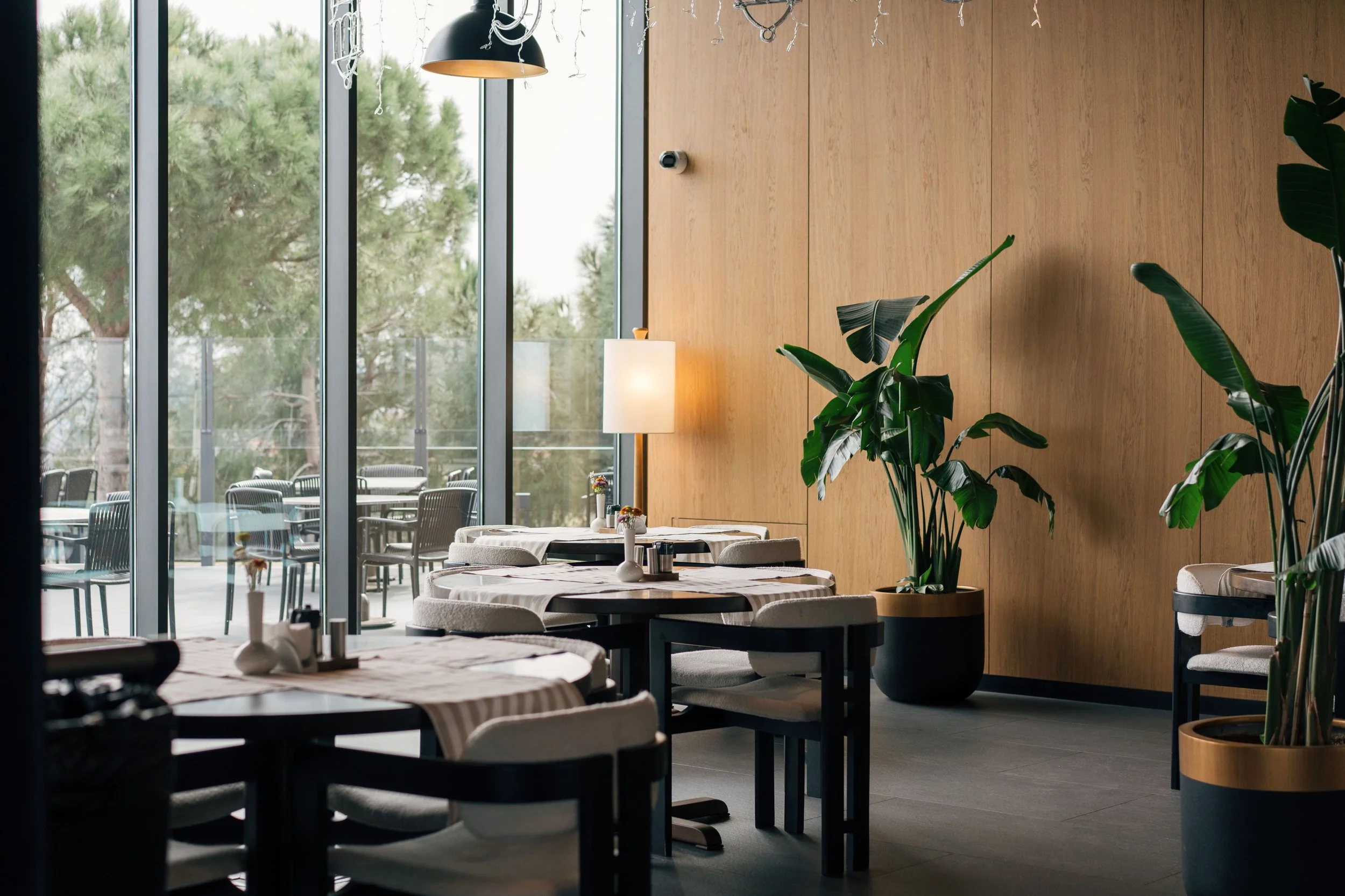Interior of a modern restaurant or cafe with large floor-to-ceiling windows, wooden wall panels, indoor plants, and tables with white tablecloths and chairs. Outside the windows are trees and an outdoor seating area.