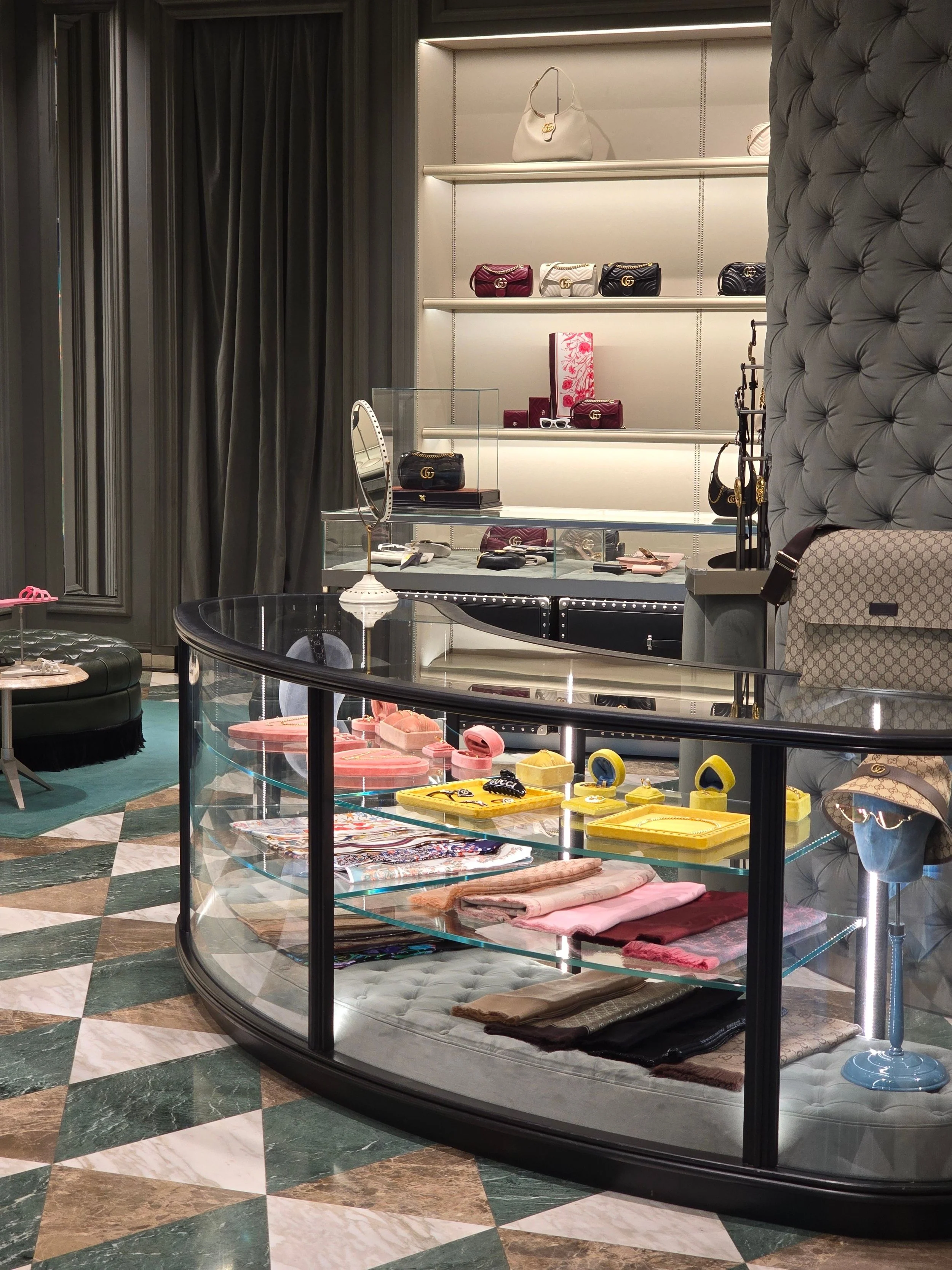 A display case in a retail store with handbags, wallets, and accessories on glass shelves. The shop has a patterned marble floor and plush seating.