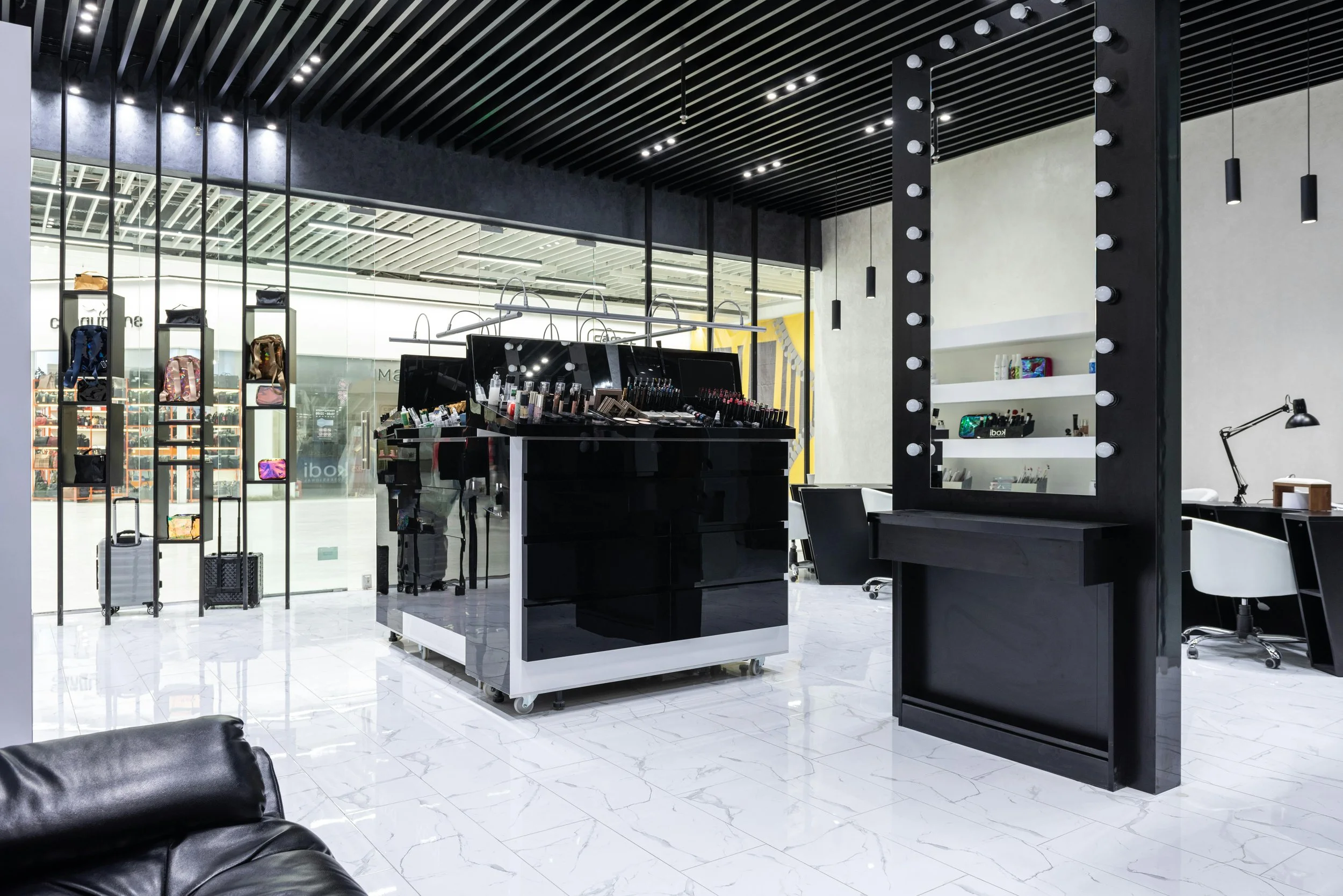 A modern makeup or cosmetic store with black and white decor, displaying makeup products on a central black counter, shelves with handbags, and large mirrors with lights, located inside a shopping mall.