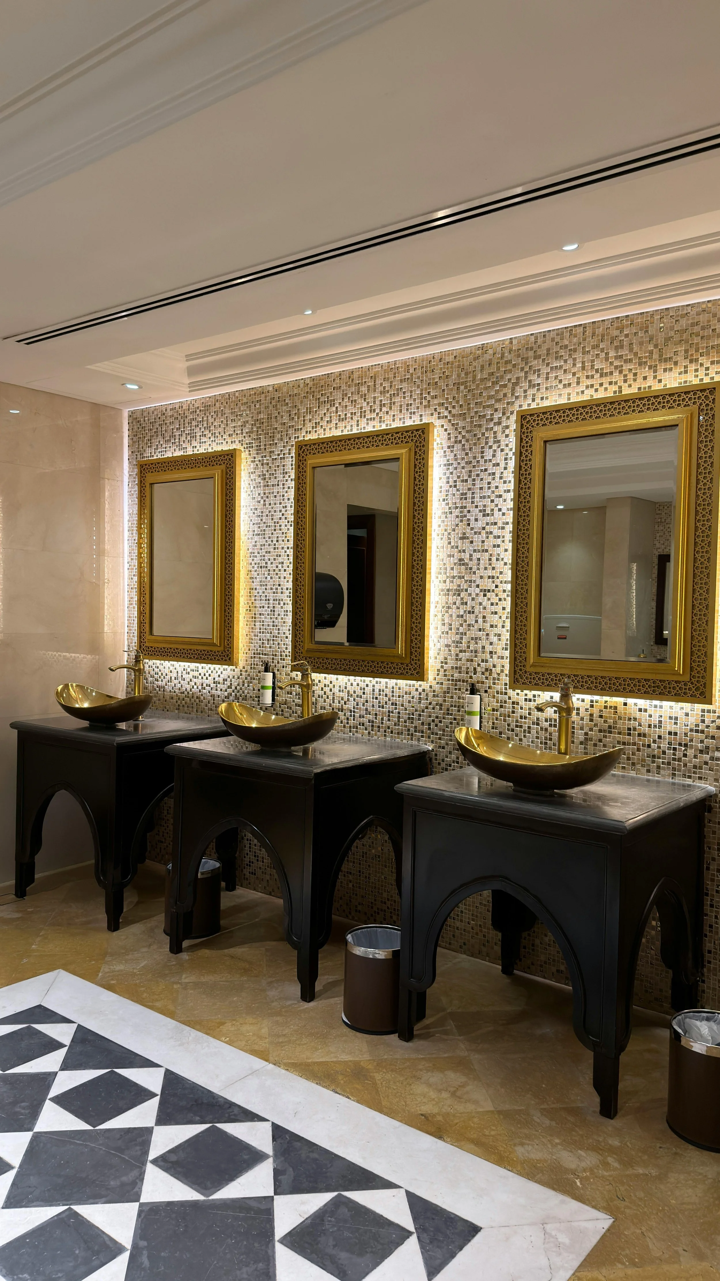 A restroom with three golden sinks on black vanities, gold-framed mirrors, a mosaic tile wall, and decorative flooring.