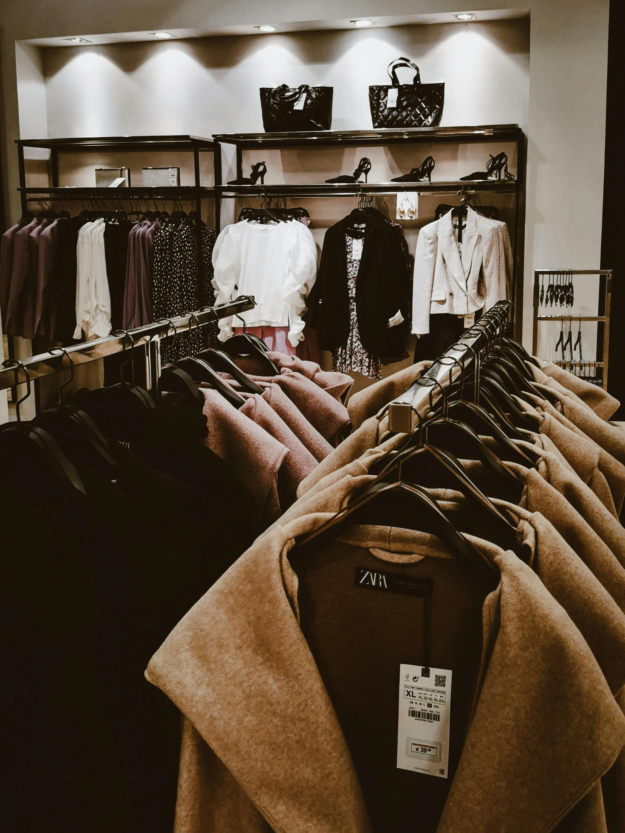 Clothing display in a store includes beige coats on hangers, with extra coats on a table in the foreground, and handbags and high heels on shelves in the background.