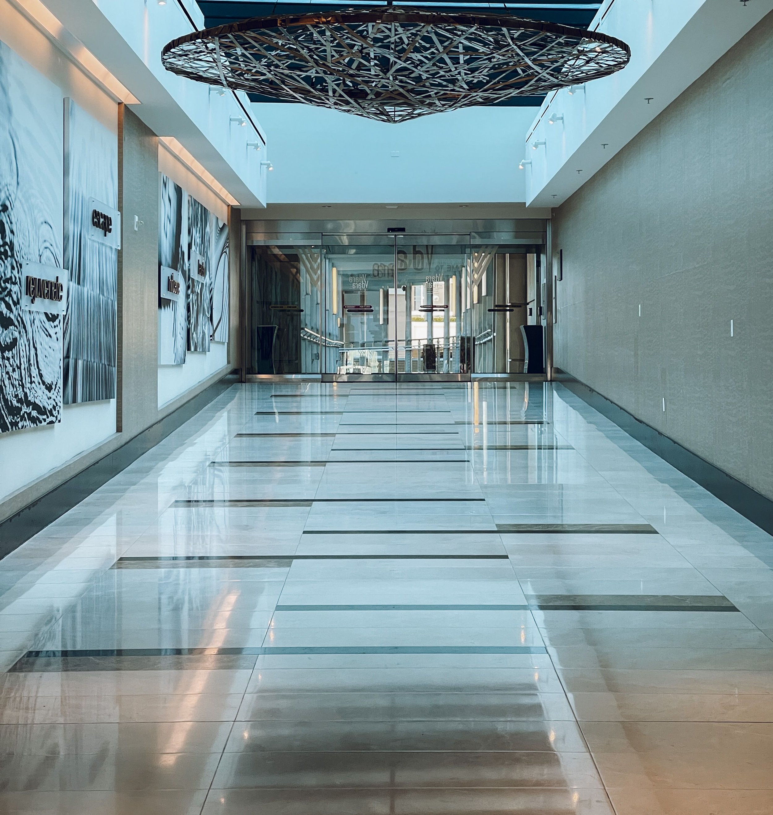 An indoor mall corridor with polished marble floors, light-colored walls, and a modern ceiling design. There is a large circular art installation hanging from the ceiling, made of interwoven wooden or metal strips. Glass doors or entrances are visible at the end of the corridor, with some signs and advertisements on the walls.