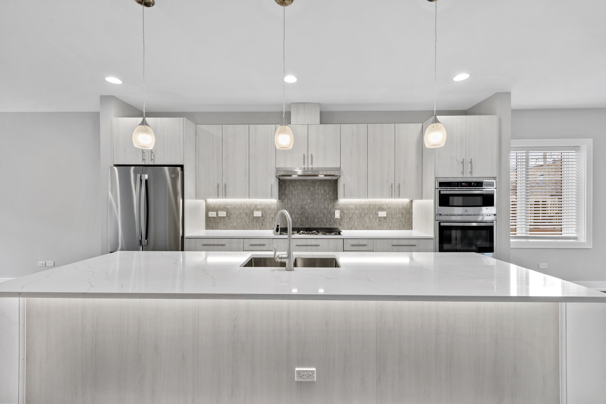 Modern kitchen with white cabinets, stainless steel appliances, pendant lights, marble island, and a window with blinds.
