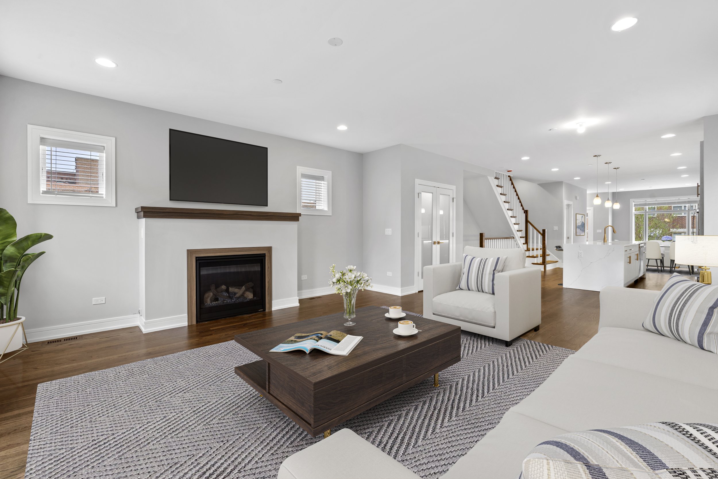 Living room with white couches, a wooden coffee table with books and cups, a fireplace, a wall-mounted TV, and a staircase leading to the upper floor, with large windows and modern decor.