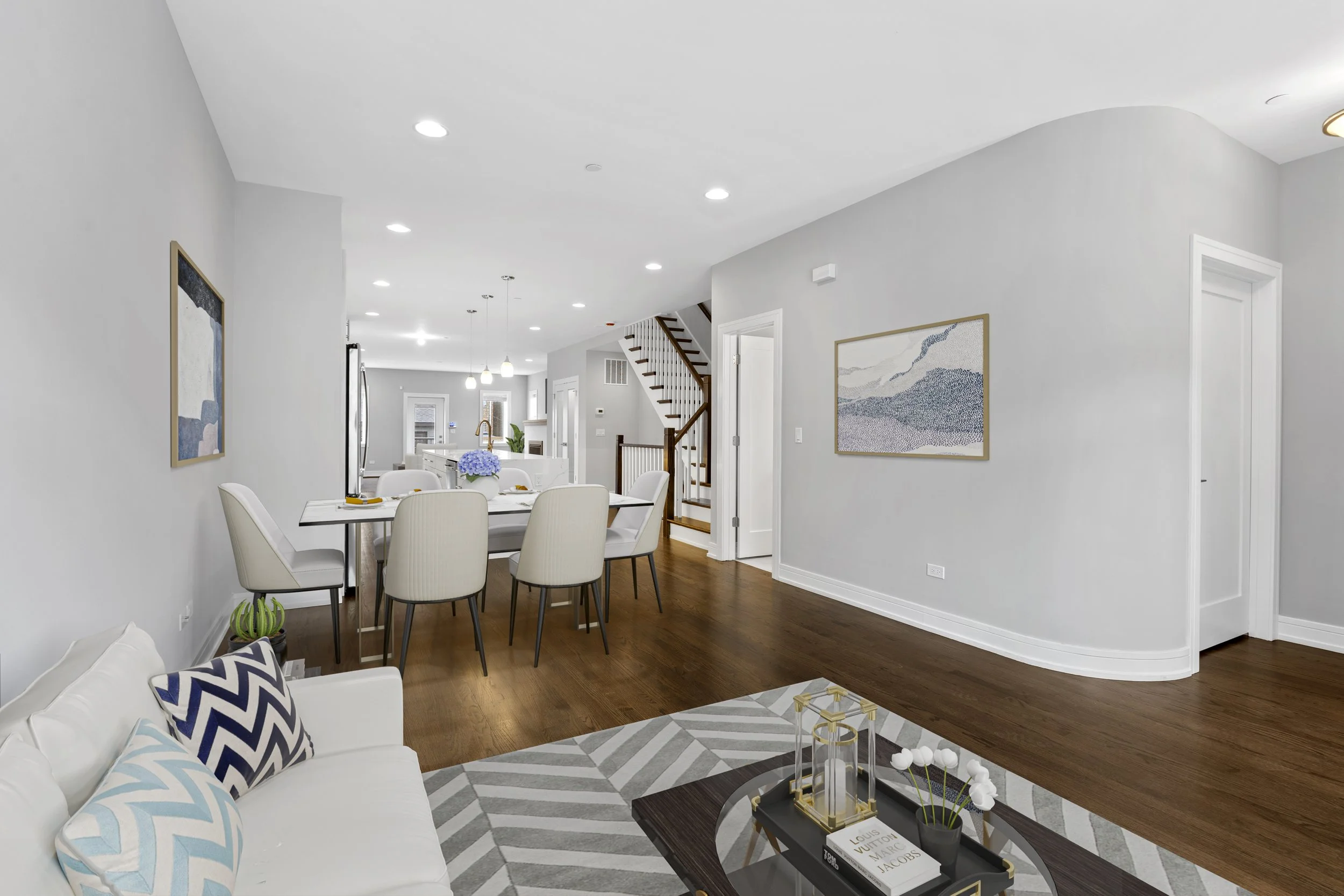 Open-concept living and dining area with white walls, hardwood floors, modern furniture, wall art, and a staircase leading upstairs.