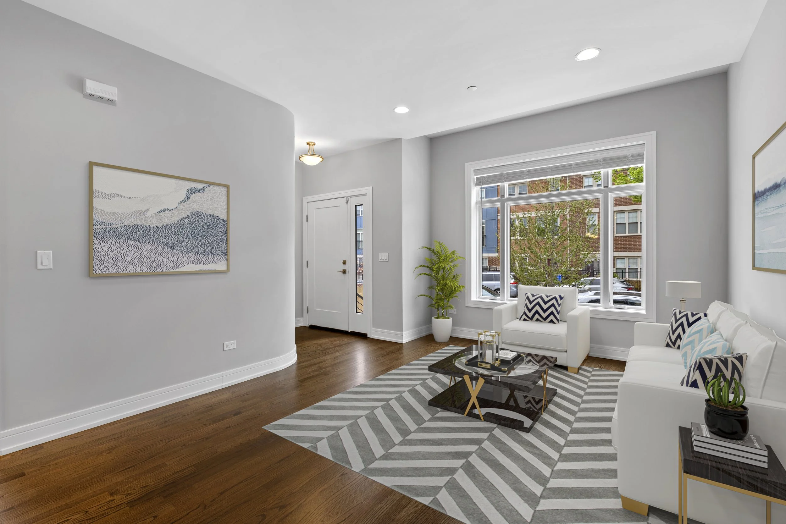 Living room with white walls, hardwood floors, large window, white furniture, striped rug, potted plants, artwork, and decorative pillows.