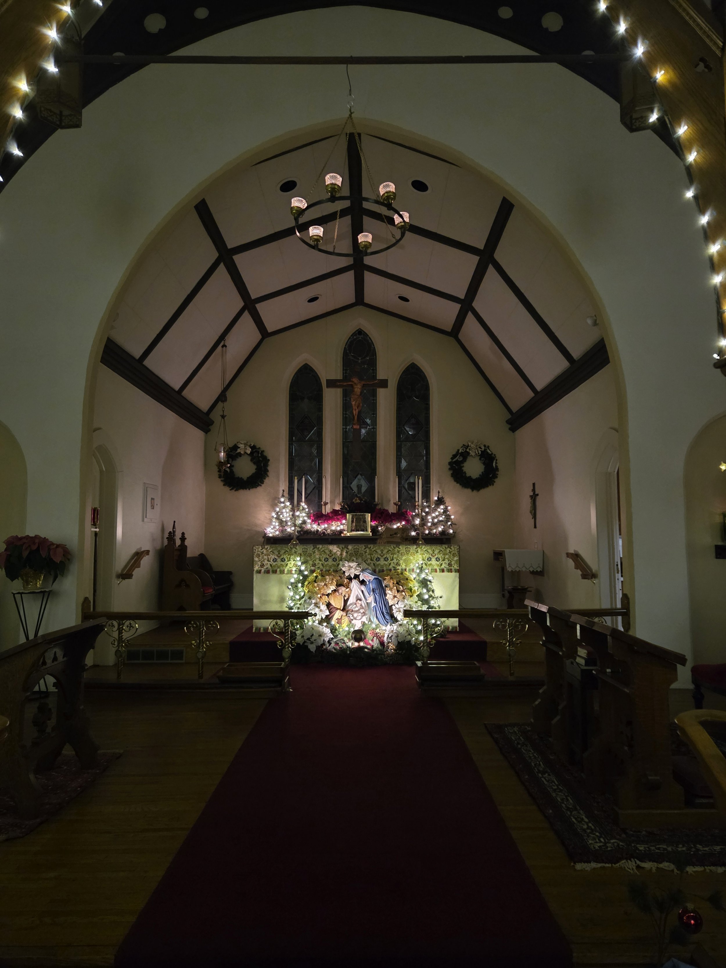 Interior of a church decorated for Christmas with wreaths, poinsettias, Christmas trees, and a nativity scene at the altar, with a crucifix and stained glass windows behind.