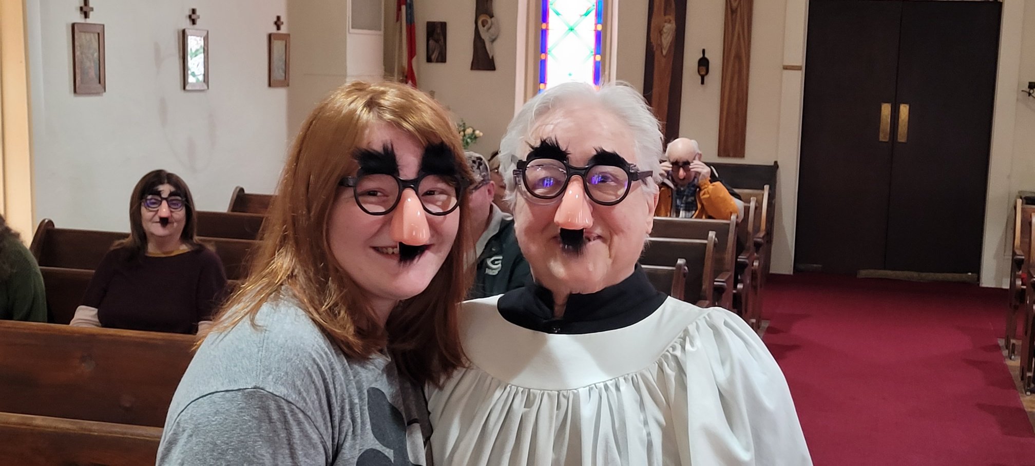 People in church pews dressed as Victorian characters with mustache and beard makeup, wearing glasses with fake noses, in a church decorated with stained glass windows and religious artwork.