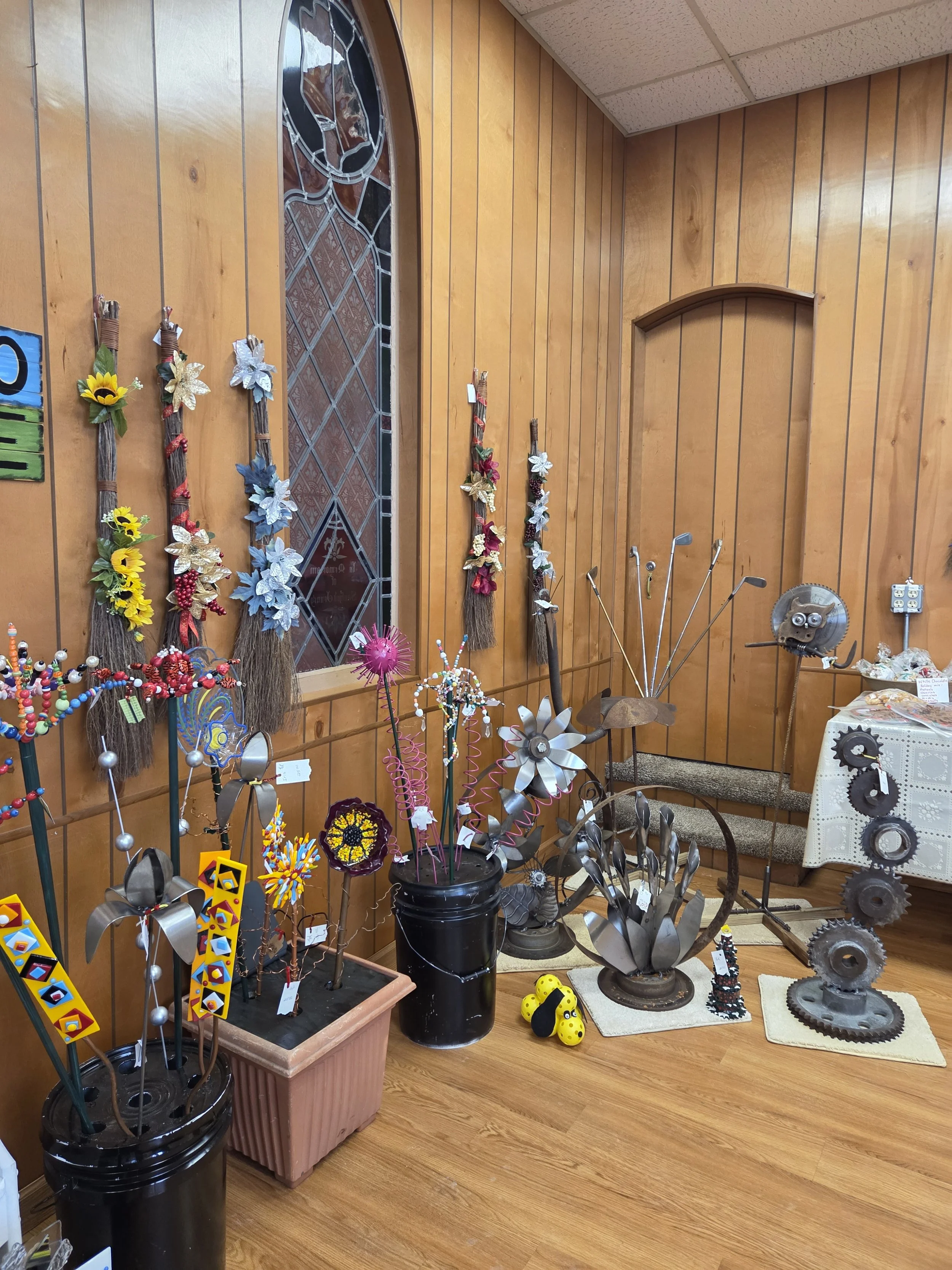 Display of handmade decorative items, including colorful metal flower sculptures, sunflower and poinsettia-like flower arrangements, and various metal gear and mechanical art pieces on tables and stands against wood-paneled walls.