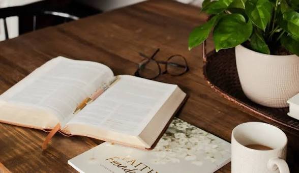 Open book, pair of reading glasses, potted plant, and a coffee mug on a wooden table.