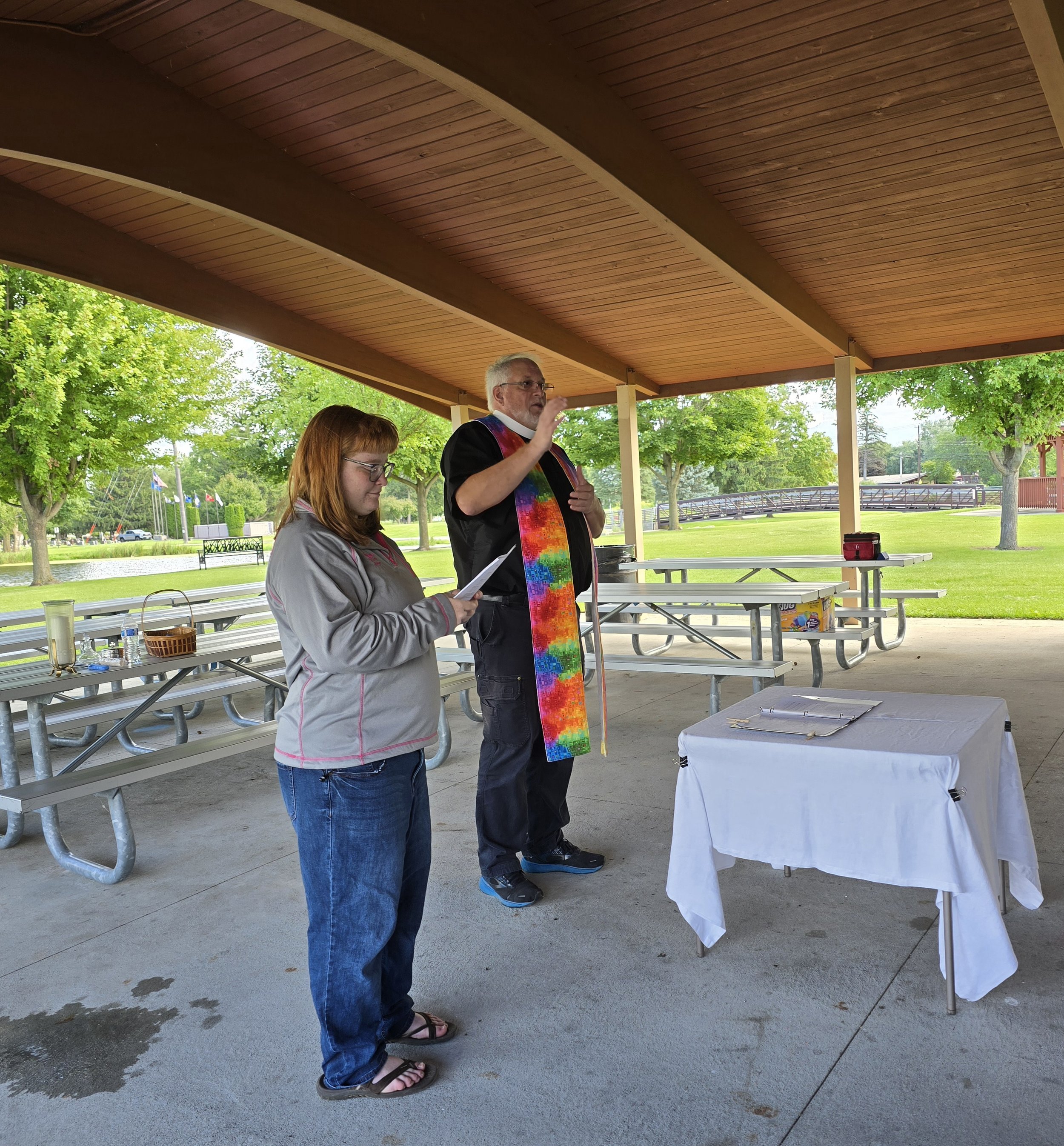 Two women and a man standing under a pavilion with wooden roof panels, participating in a gathering or ceremony in an outdoor park with trees and benches in the background. The man is wearing glasses and a rainbow sash, speaking, while one woman is h