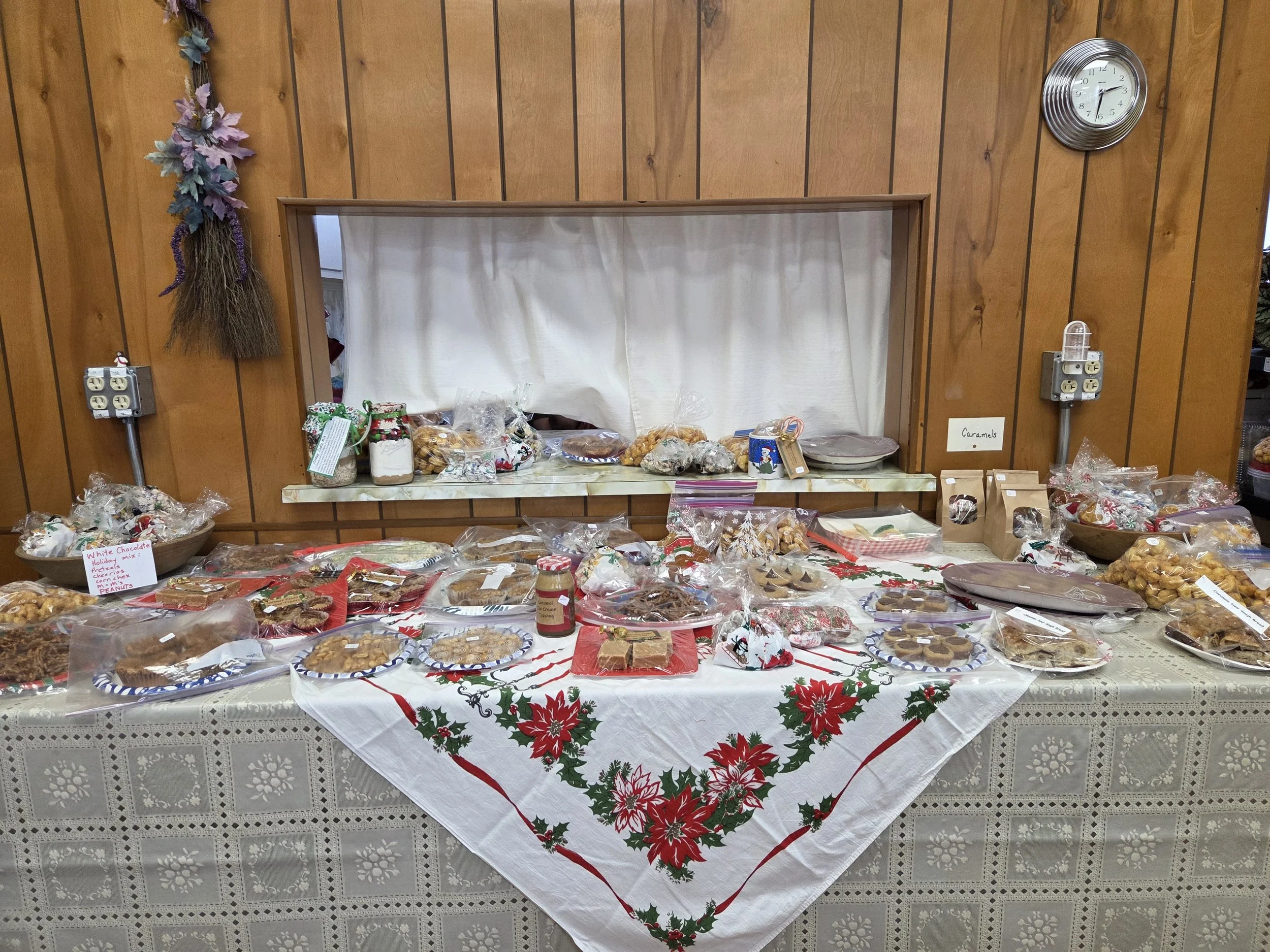A holiday-themed table with various baked goods and treats, spread on a table covered with a holiday cloth. The table is set against a wood-paneled wall with a small window, decorated with a hanging floral arrangement and a clock.