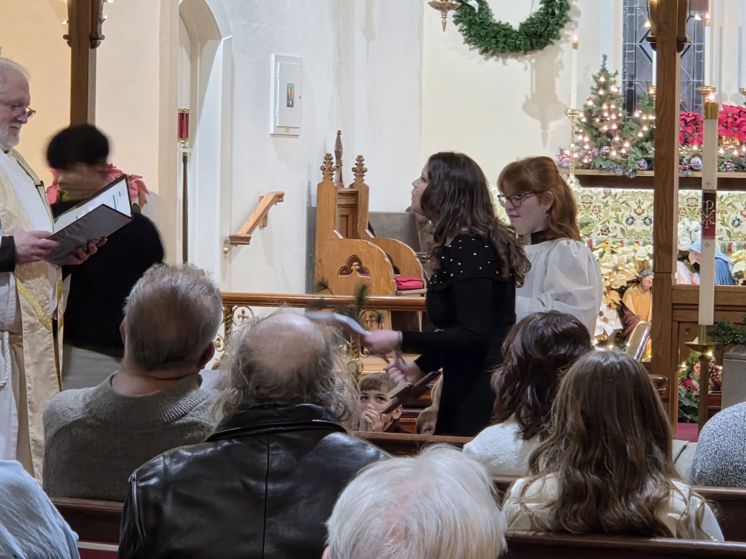 People gathered inside a church decorated for Christmas, with a Christmas tree and wreaths, as a choir performs.