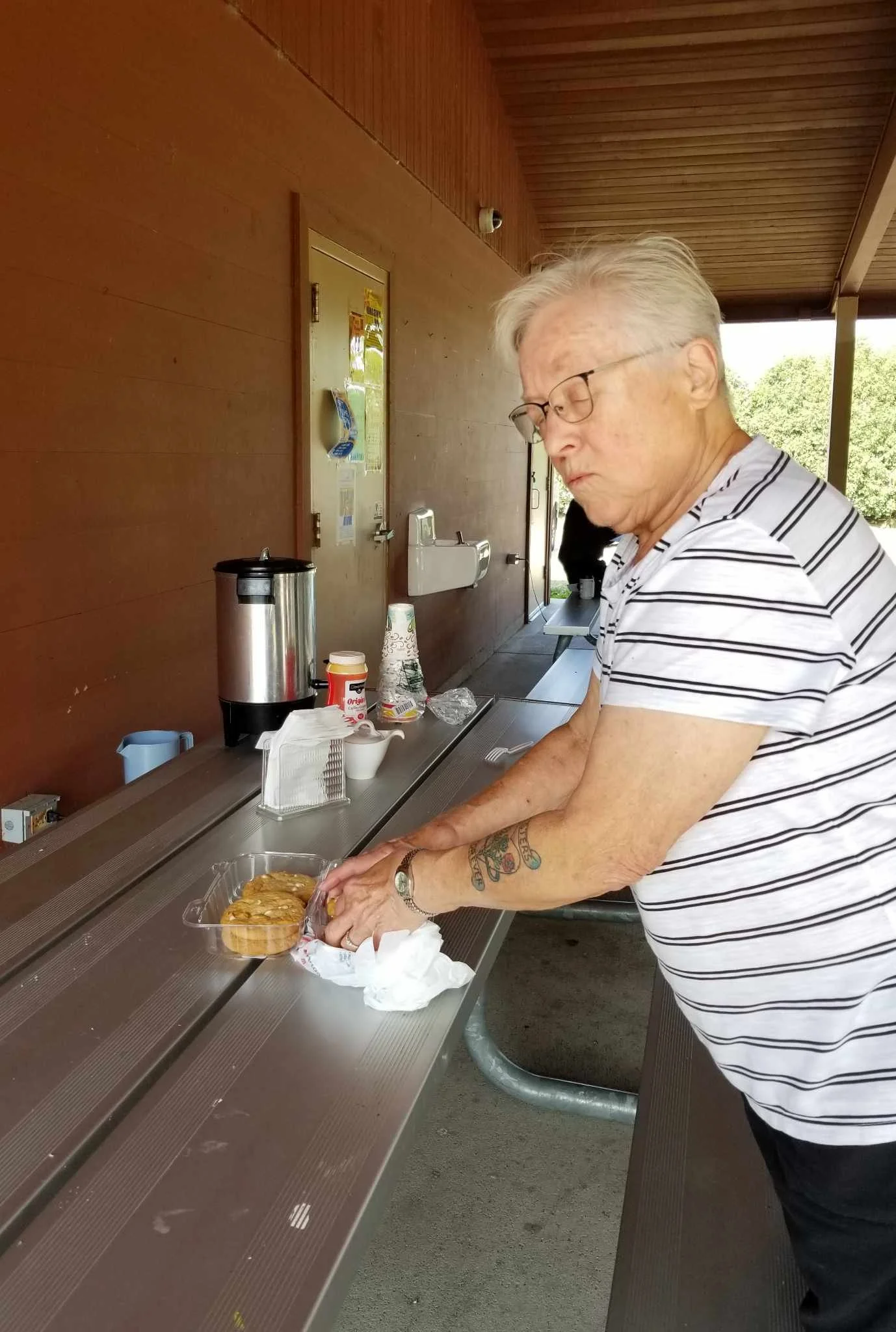 Older man with gray hair and glasses wearing a striped shirt, preparing food at an outdoor picnic table. There are snacks, cups, and a travel mug on the table, and a wooden wall with a paper towel dispenser in the background.