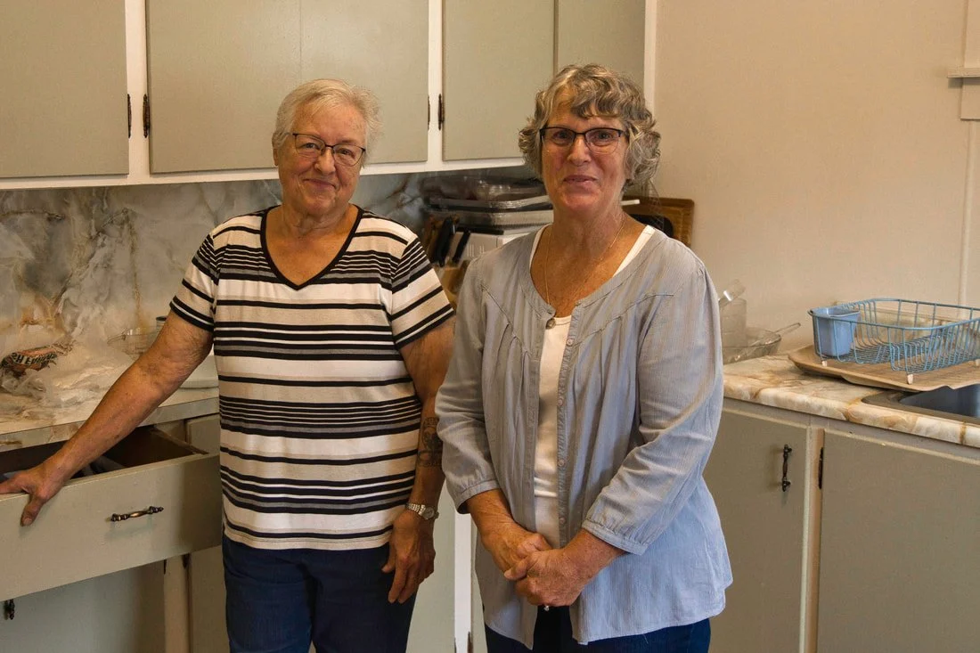 Two older women standing in a kitchen, smiling at the camera. One woman on the left wears a black and white striped shirt, and the other on the right wears glasses and a light gray jacket.
