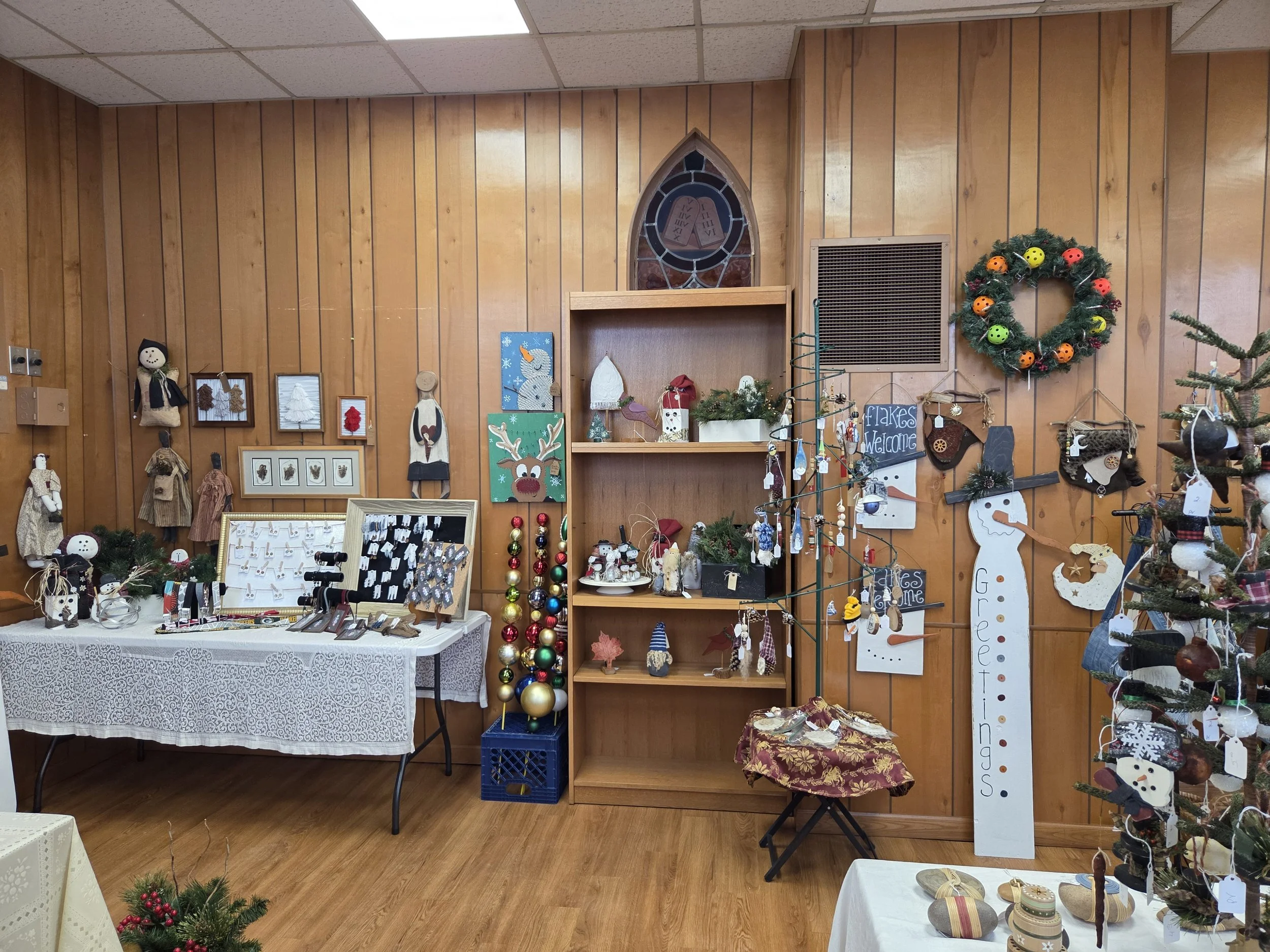 A room decorated with Christmas and winter holiday themed ornaments and crafts. There are wooden wall panels, a stained glass window, and various holiday decorations including a wreath with colorful ornaments, crafted snowmen, reindeer, and other fes