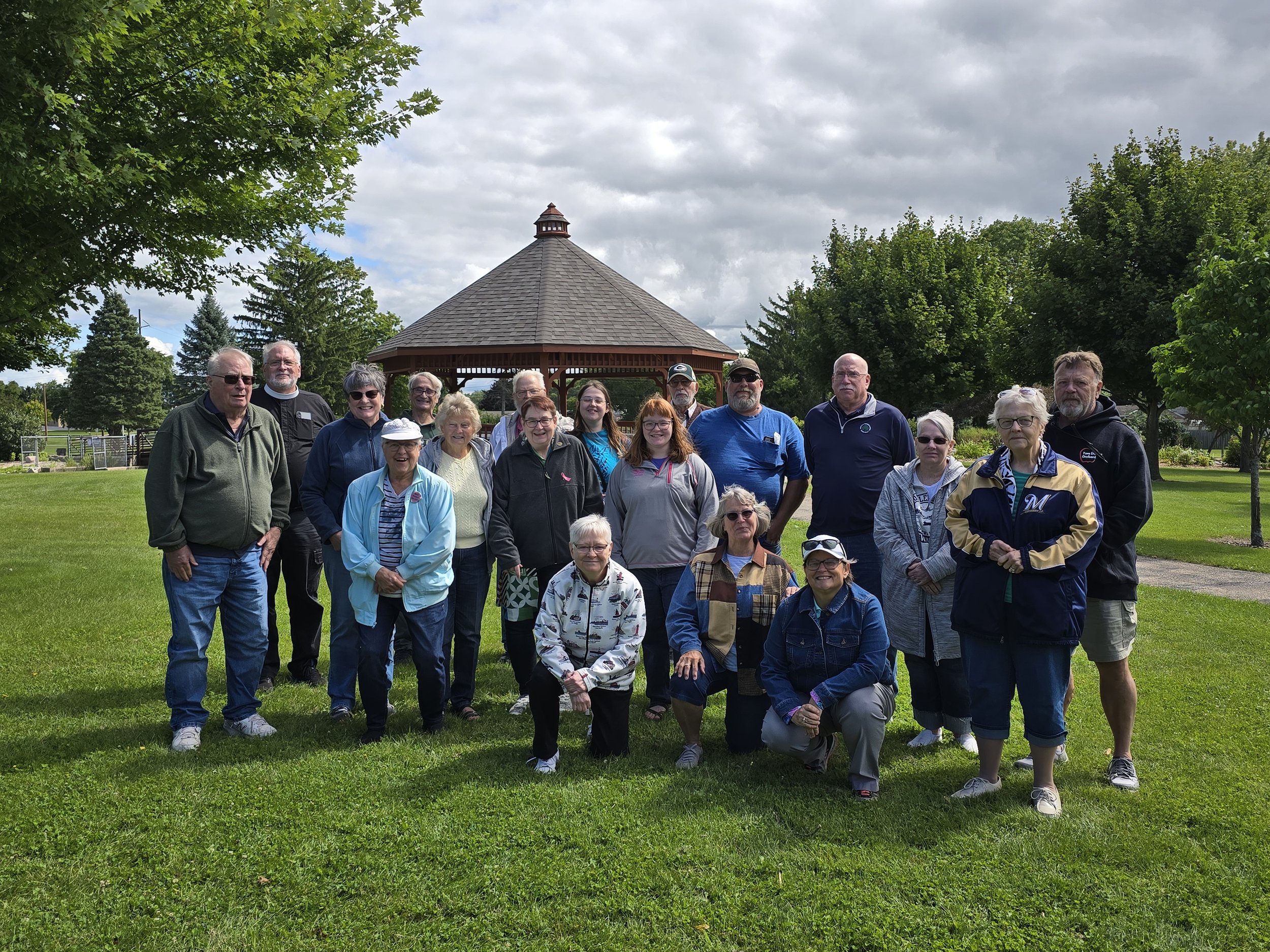 A large group of people, mostly seniors, standing outdoors in front of a gazebo in a park on a cloudy day.