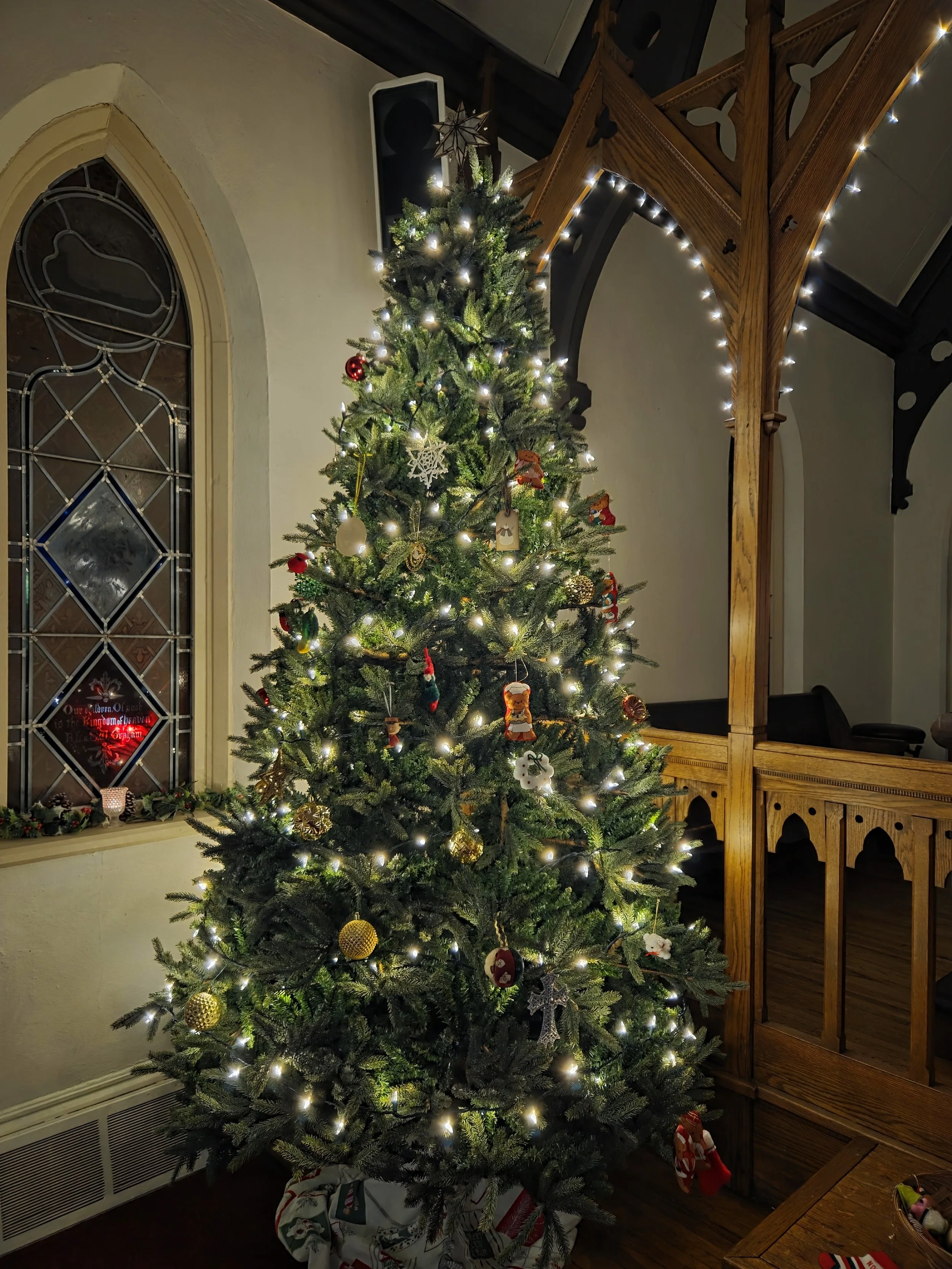 A decorated Christmas tree with lights and ornaments inside a house near a window and wooden railing.