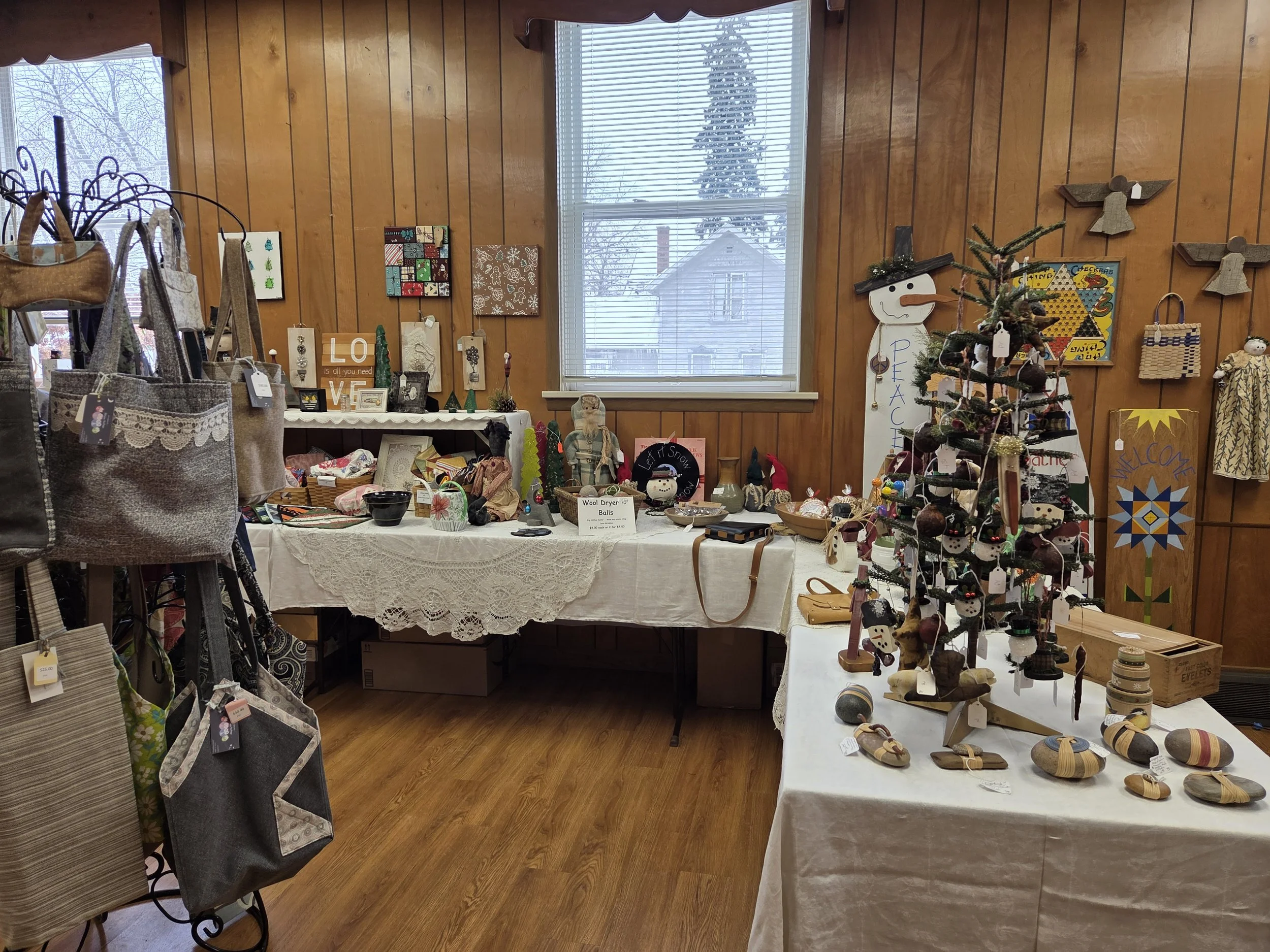 A display of various holiday-themed craft items and decorations on tables in a wood-paneled room with a window showing a snowy outdoor scene.