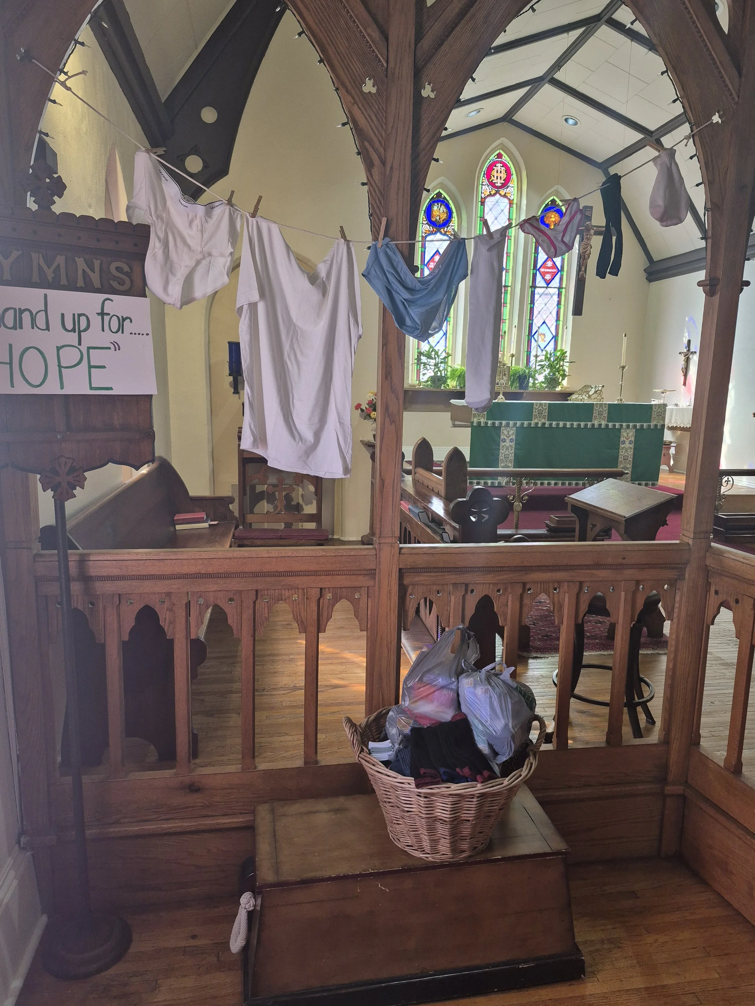 A wooden church booth with a hanging laundry line with white, blue, and black clothing. Inside the church, there are stained glass windows, a green altar cloth, and a crucifix on the wall. There is a basket of clothes on the floor and a small wooden step.