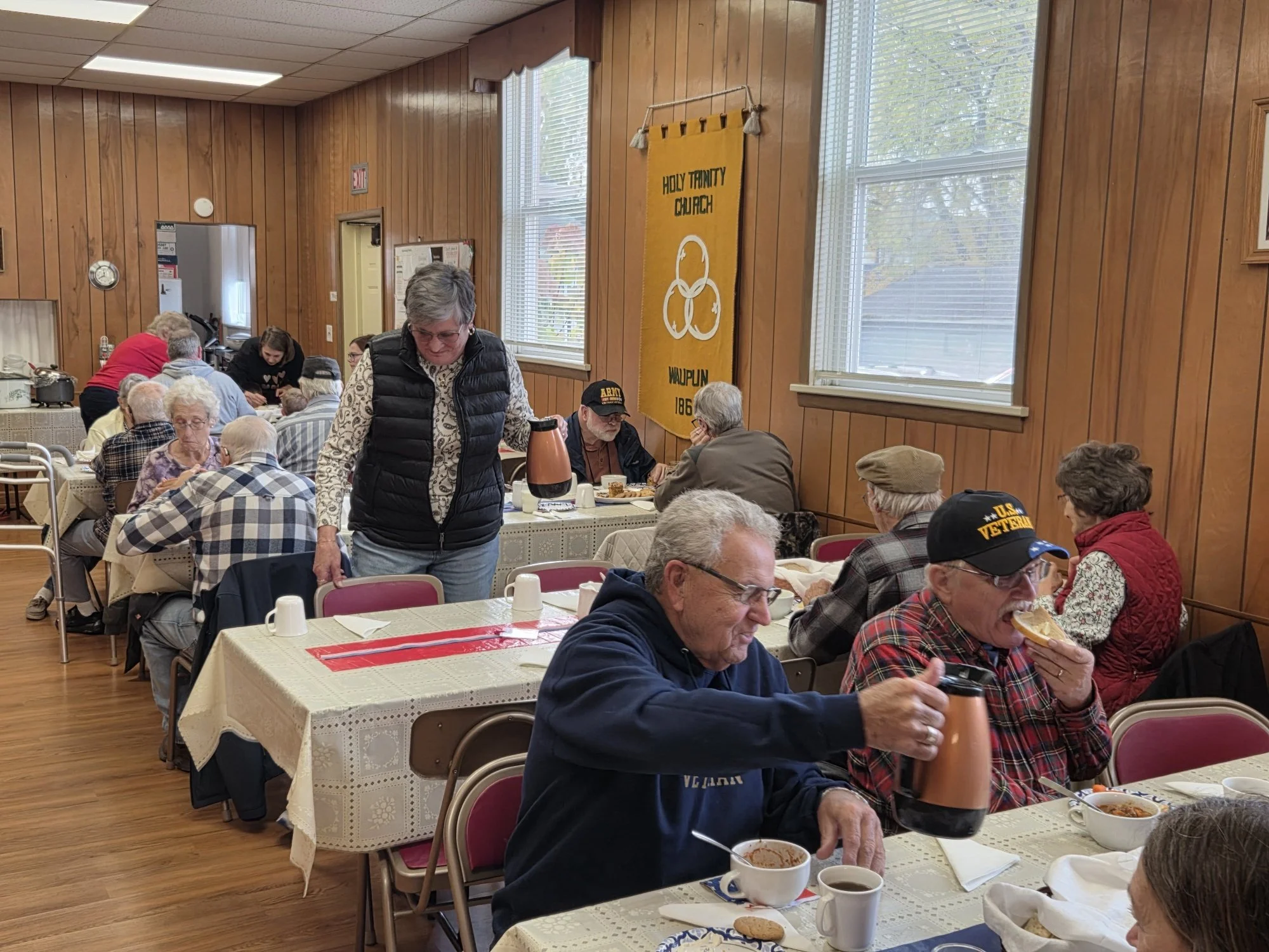 A group of elderly people dining together in a Wood-paneled community hall, with some people eating and others sitting and talking. A woman with glasses and a black vest is serving coffee, and there are banners and windows in the background.