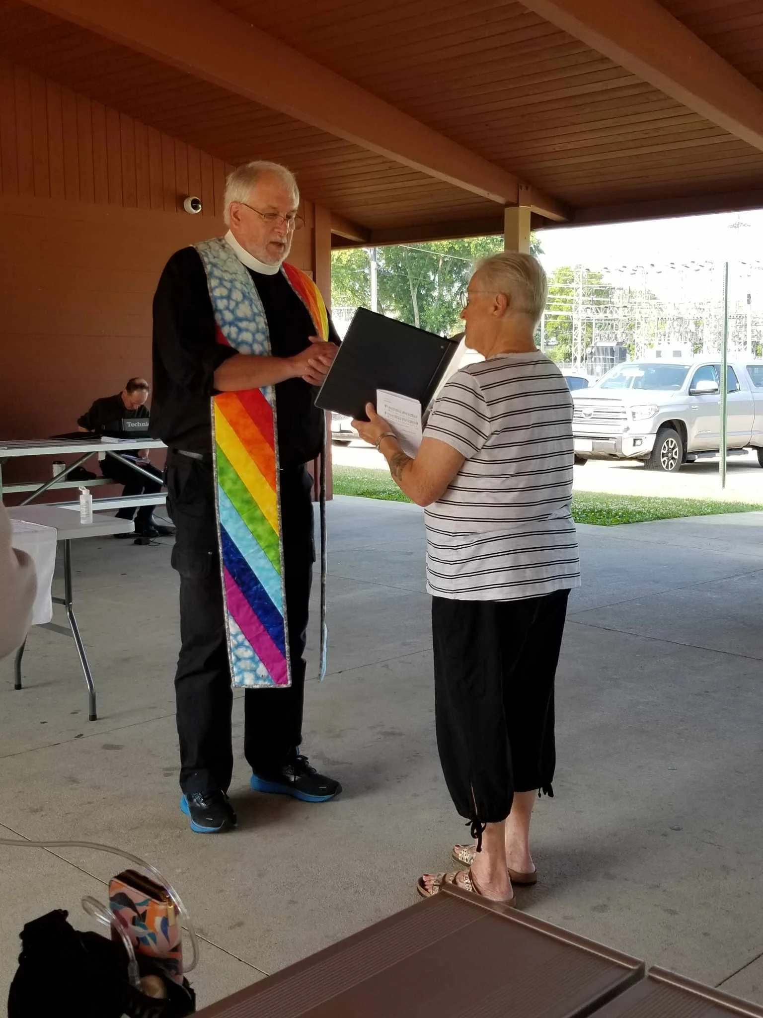 A man with glasses presents an award or certificate to a woman with short, light-colored hair at an outdoor event under a covered pavilion. Another person is sitting at a table in the background.