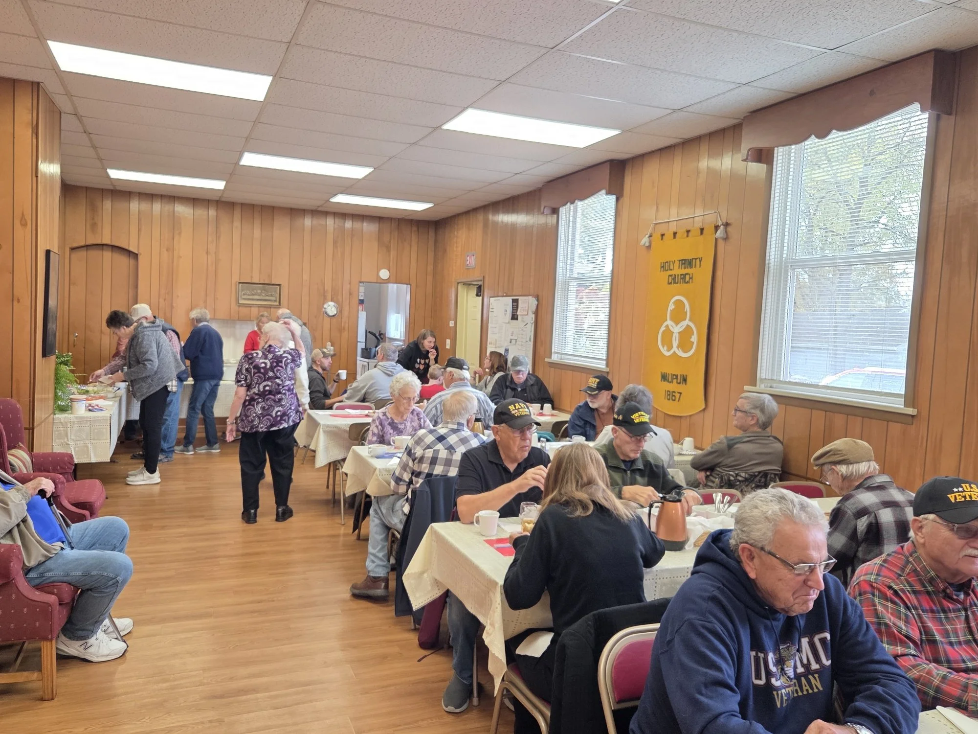 Veterans gathering in a community hall for a meal, some are seated and some are standing near a table with food.