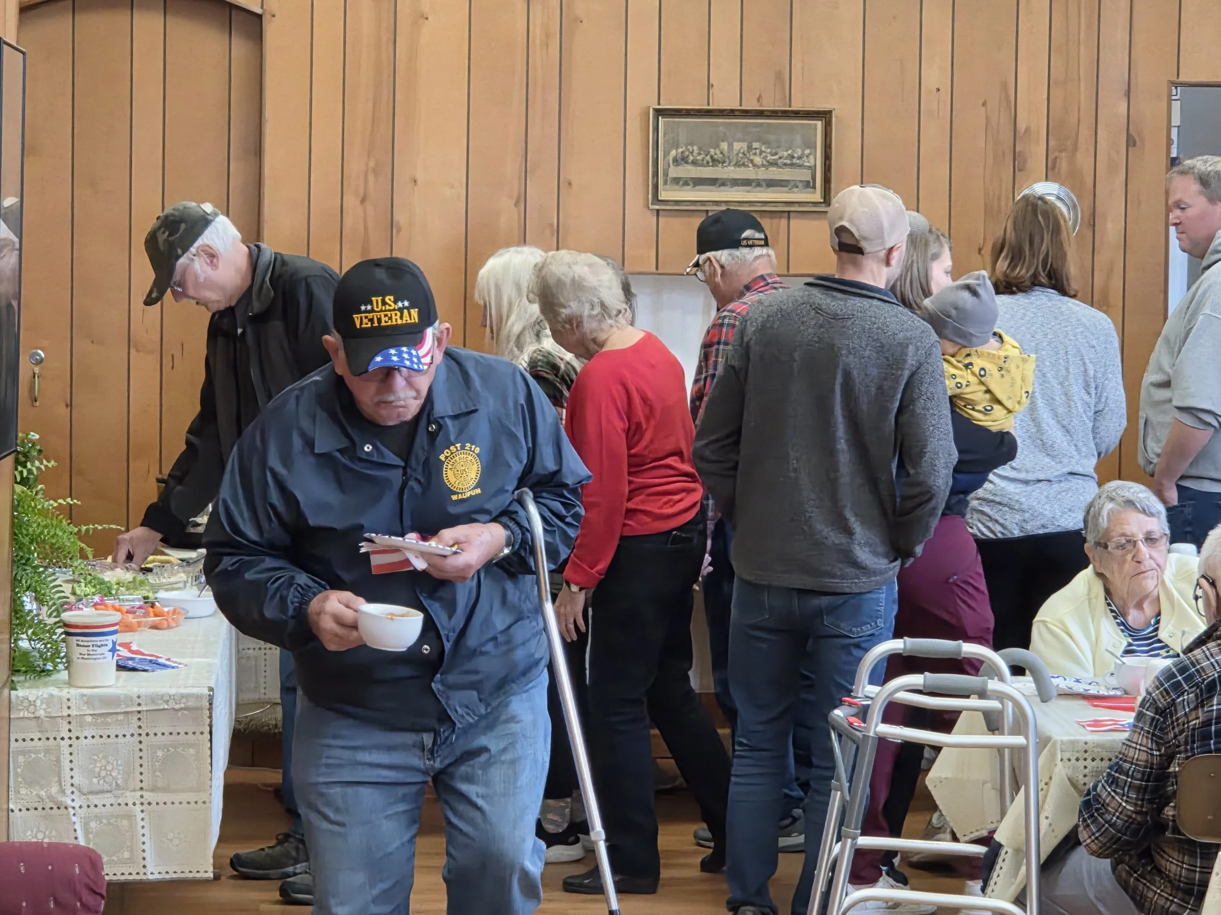 People standing in line at a cafeteria table for food, with some seated and eating. The setting appears to be a community center or senior living facility with wood-paneled walls and framed artwork. The individuals include elderly and middle-aged people, some wearing hats, and one man with a U.S. veteran cap.