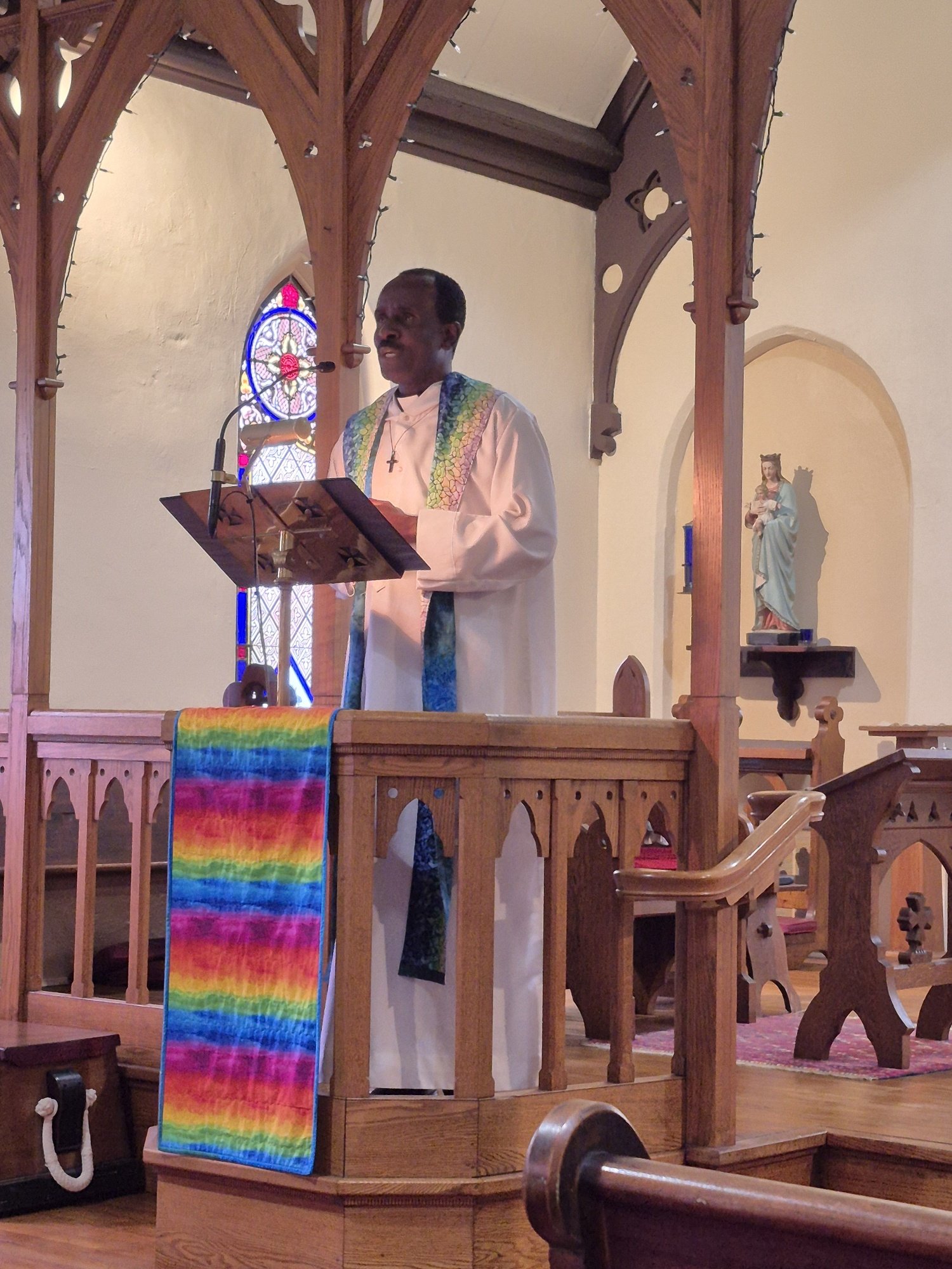 A man dressed in white with a cross necklace standing at a wooden lectern inside a church, with a stained glass window and religious statue in the background.