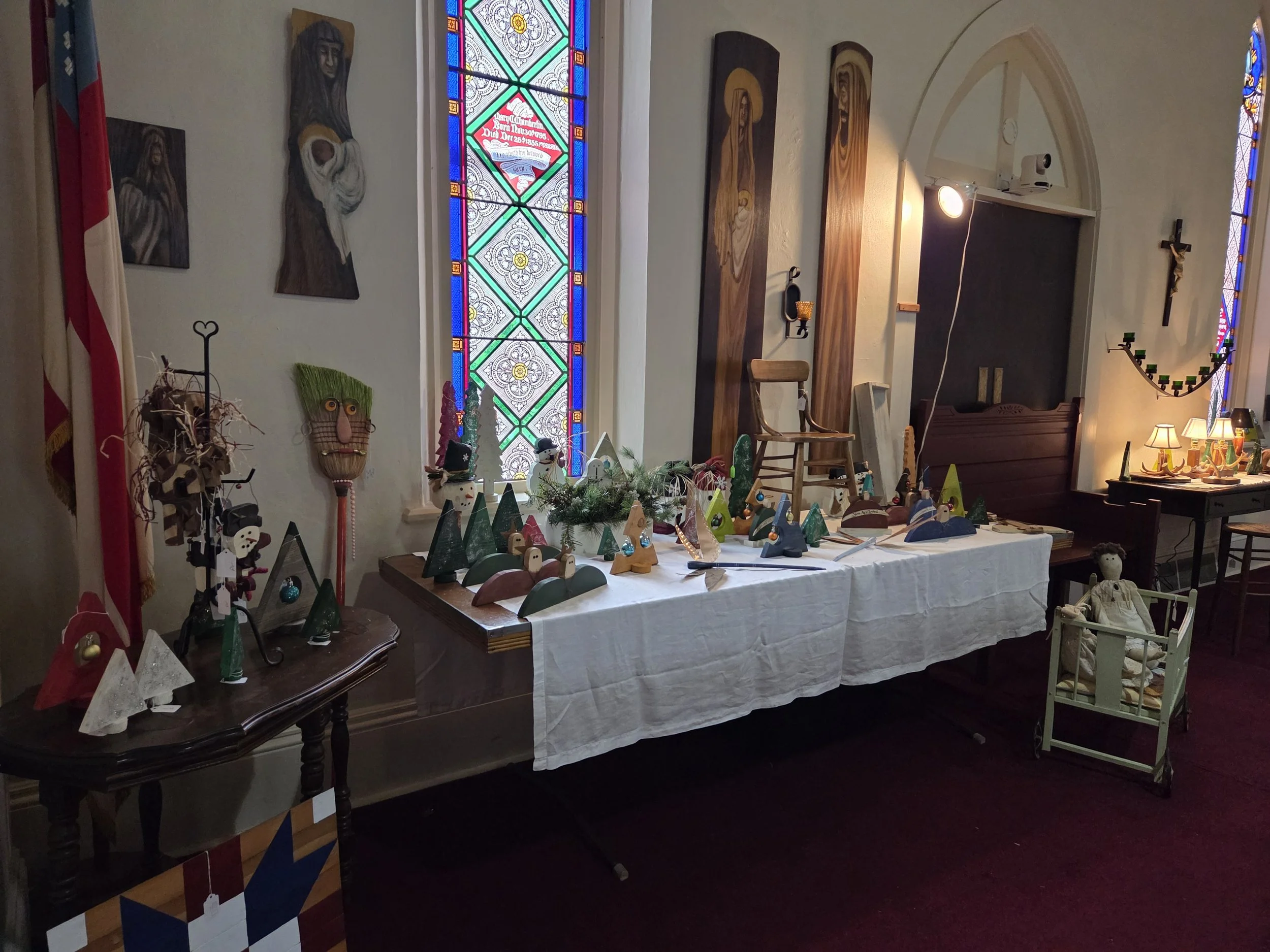Indoor church scene with stained glass windows, religious artwork, a table displaying Christmas crafts, candles, and a crucifix on the wall.