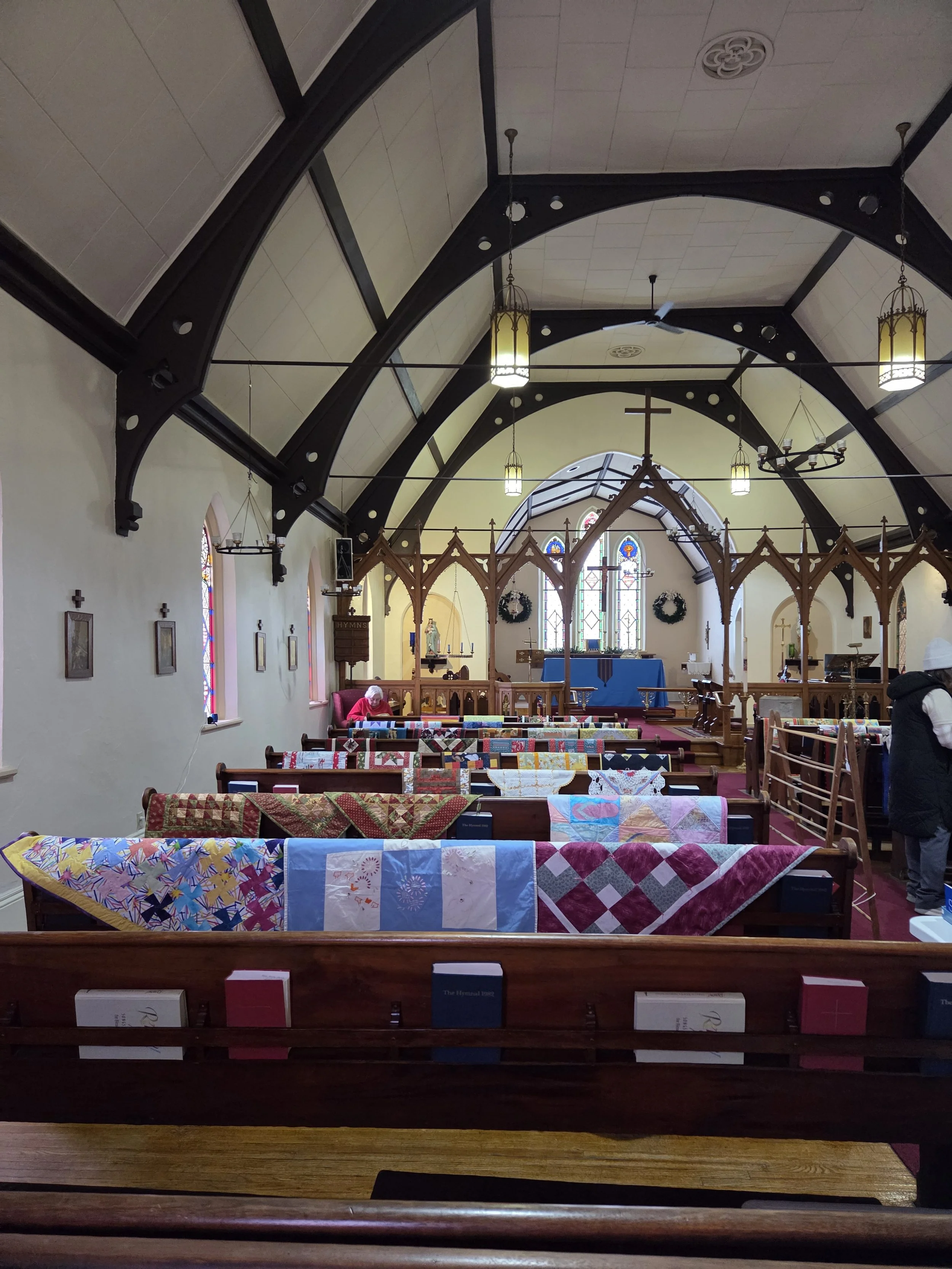Interior of a church with wooden pews adorned with colorful and patterned quilts, stained glass windows, and religious symbols including a cross and statues, lit by hanging lamps.