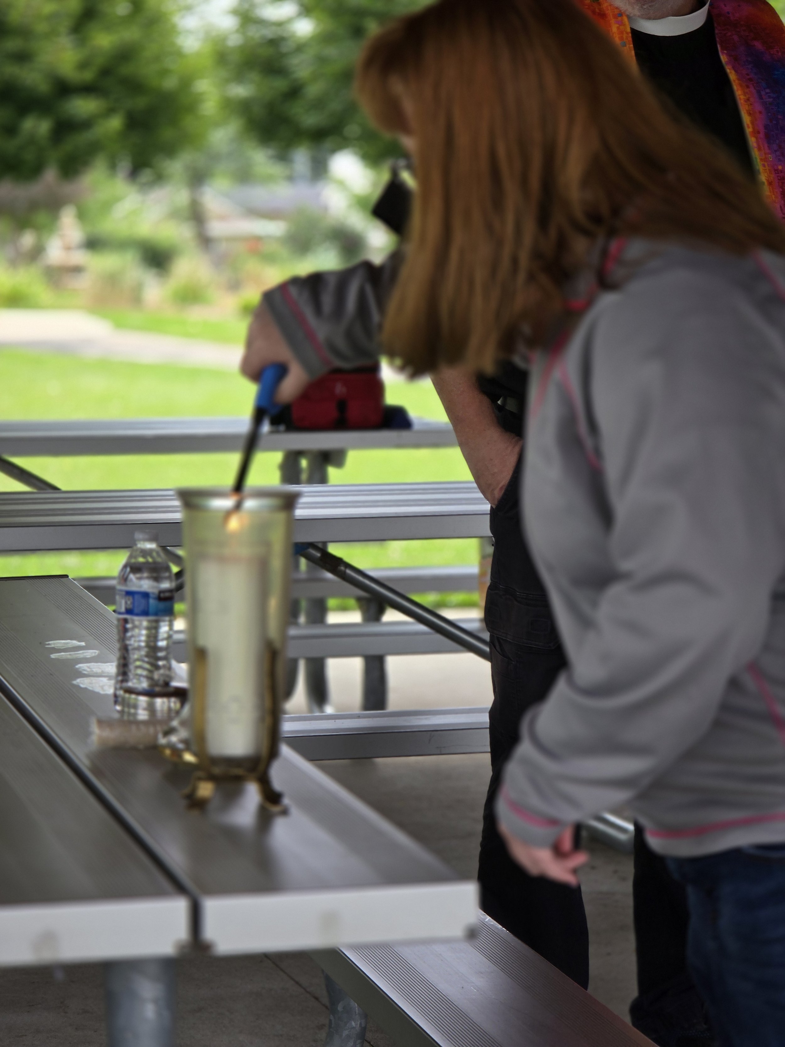 People standing at a stainless steel outdoor food serving counter with beverages, in a park setting with green trees in the background.