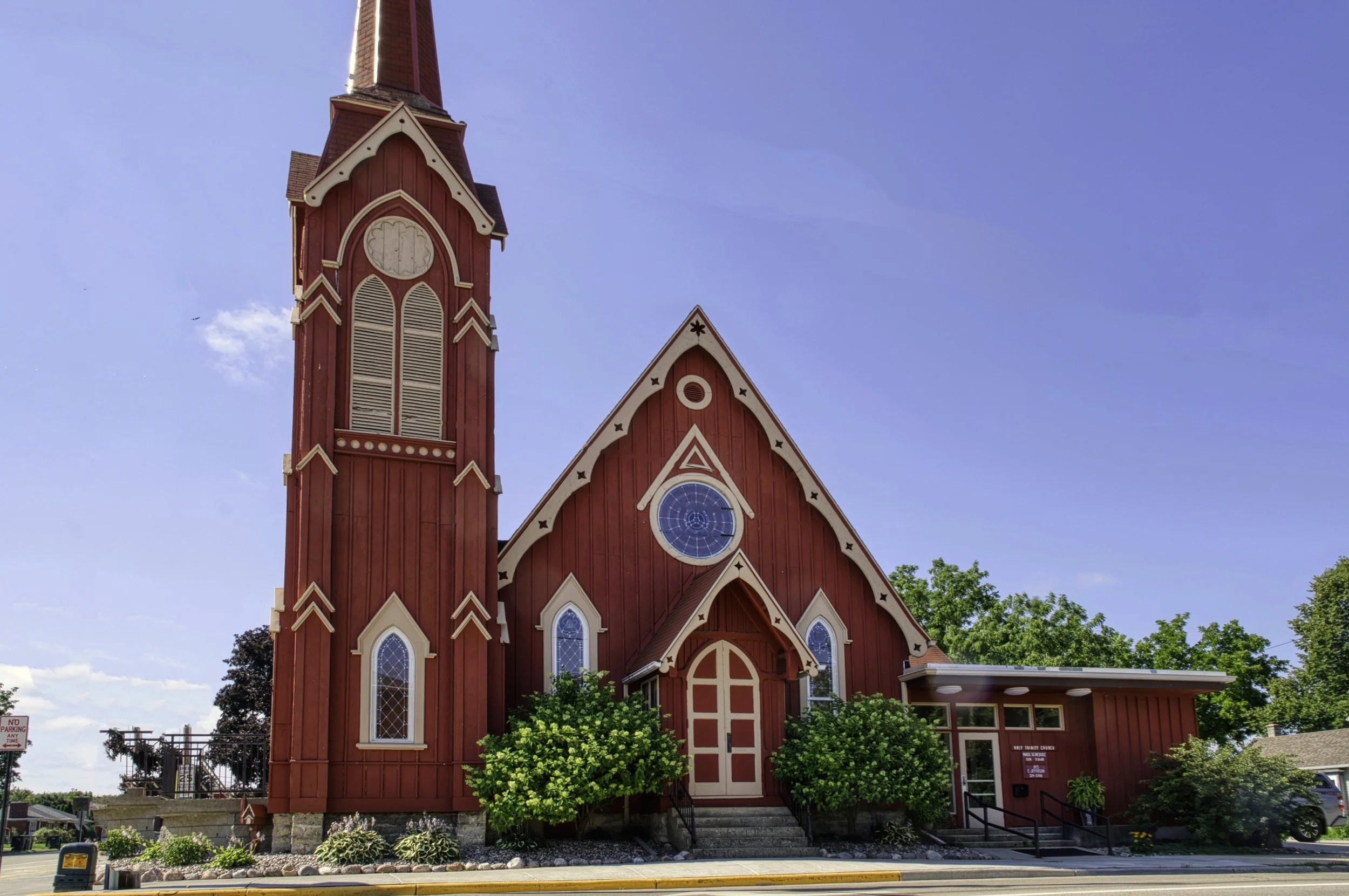 Red wooden church with tall steeple and stained glass windows, surrounded by greenery and a sidewalk, under a blue sky.