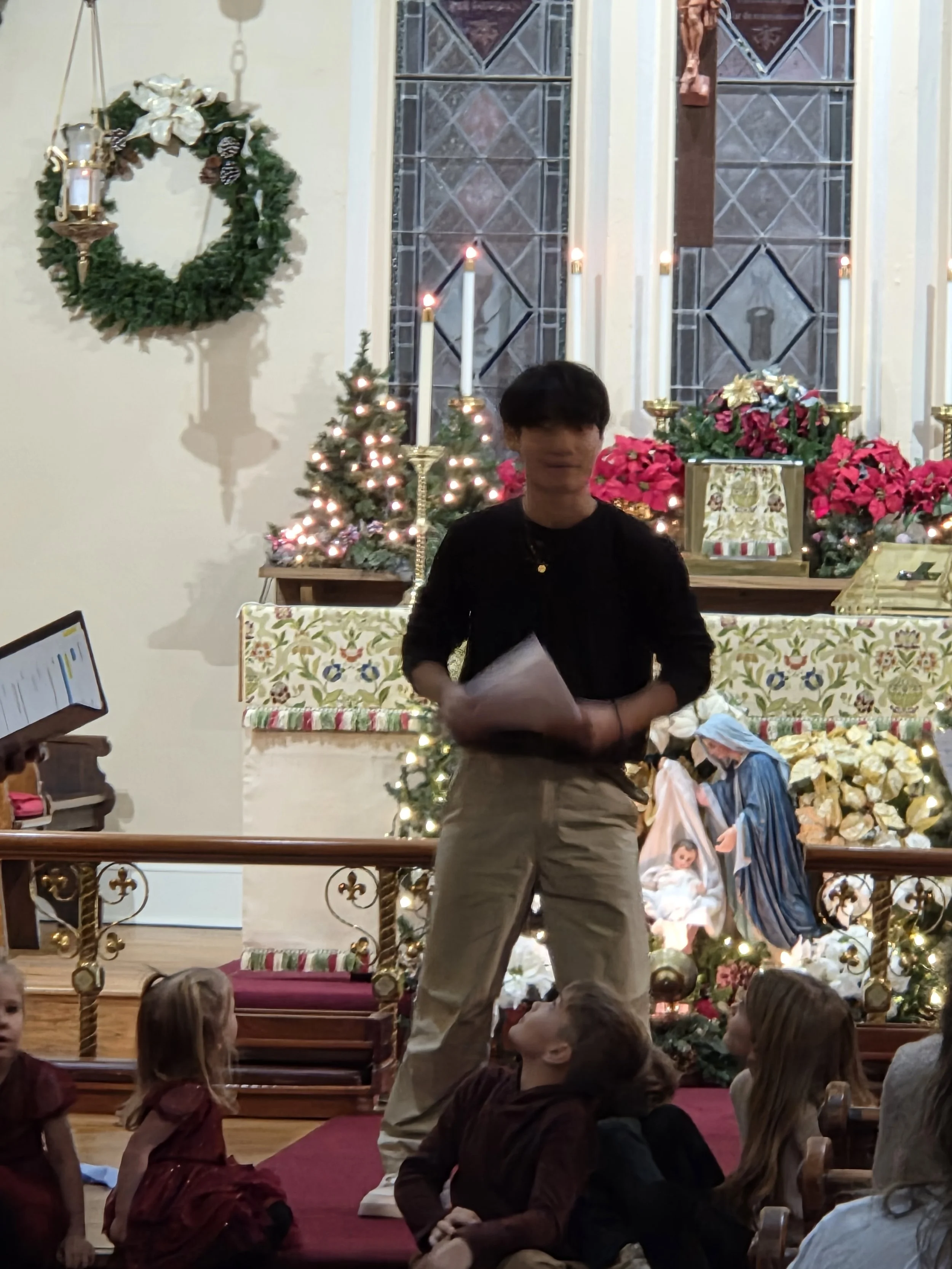 A nativity scene inside a church with children and an adult. The background decorated with Christmas plants, candles, and a manger scene with figures of Mary, Joseph, and baby Jesus.