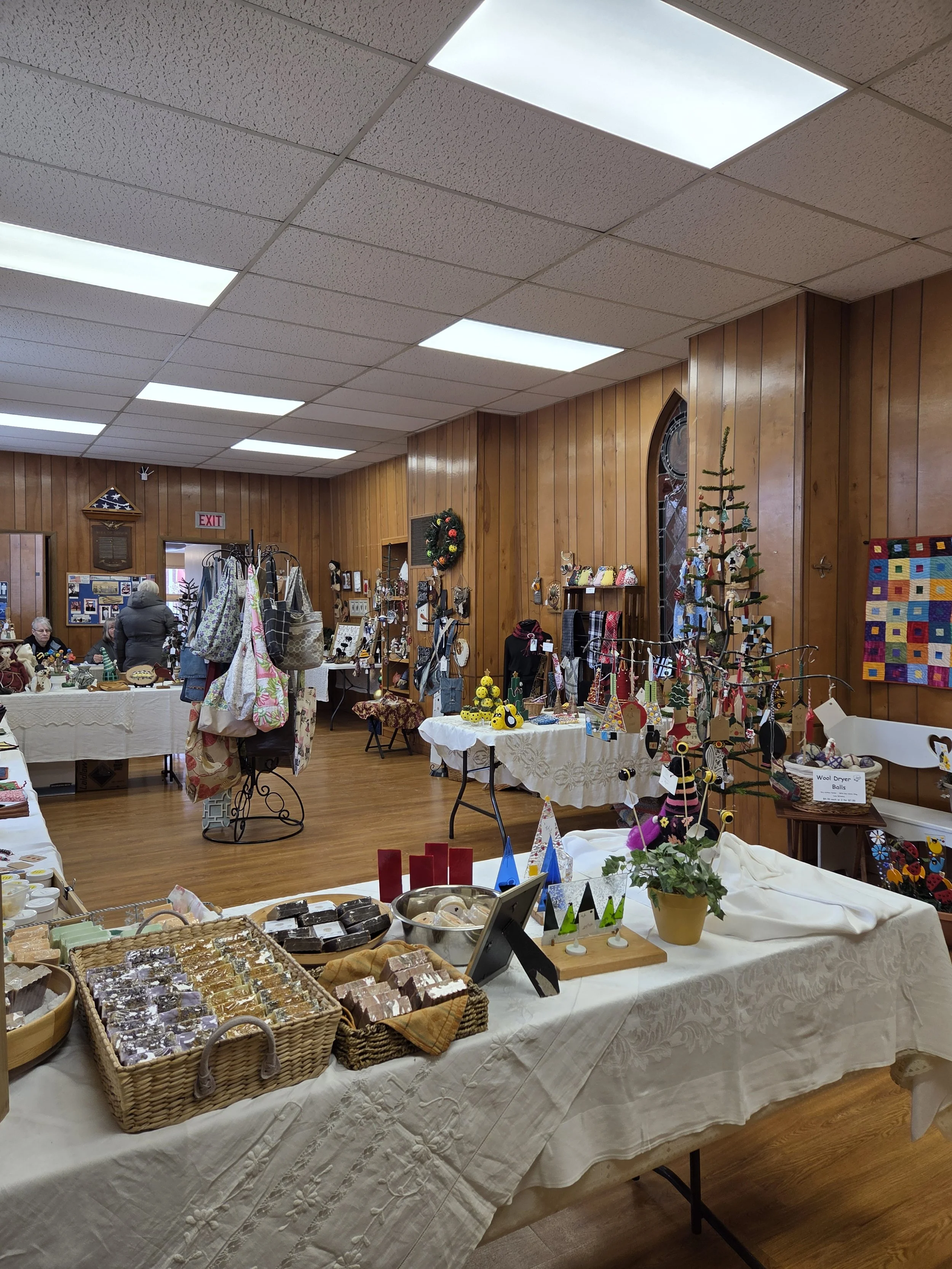 Indoor craft show displaying handmade items such as jewelry, decorations, and baked goods on tables covered with white tablecloths, with people browsing and wooden wall paneling inside the room.
