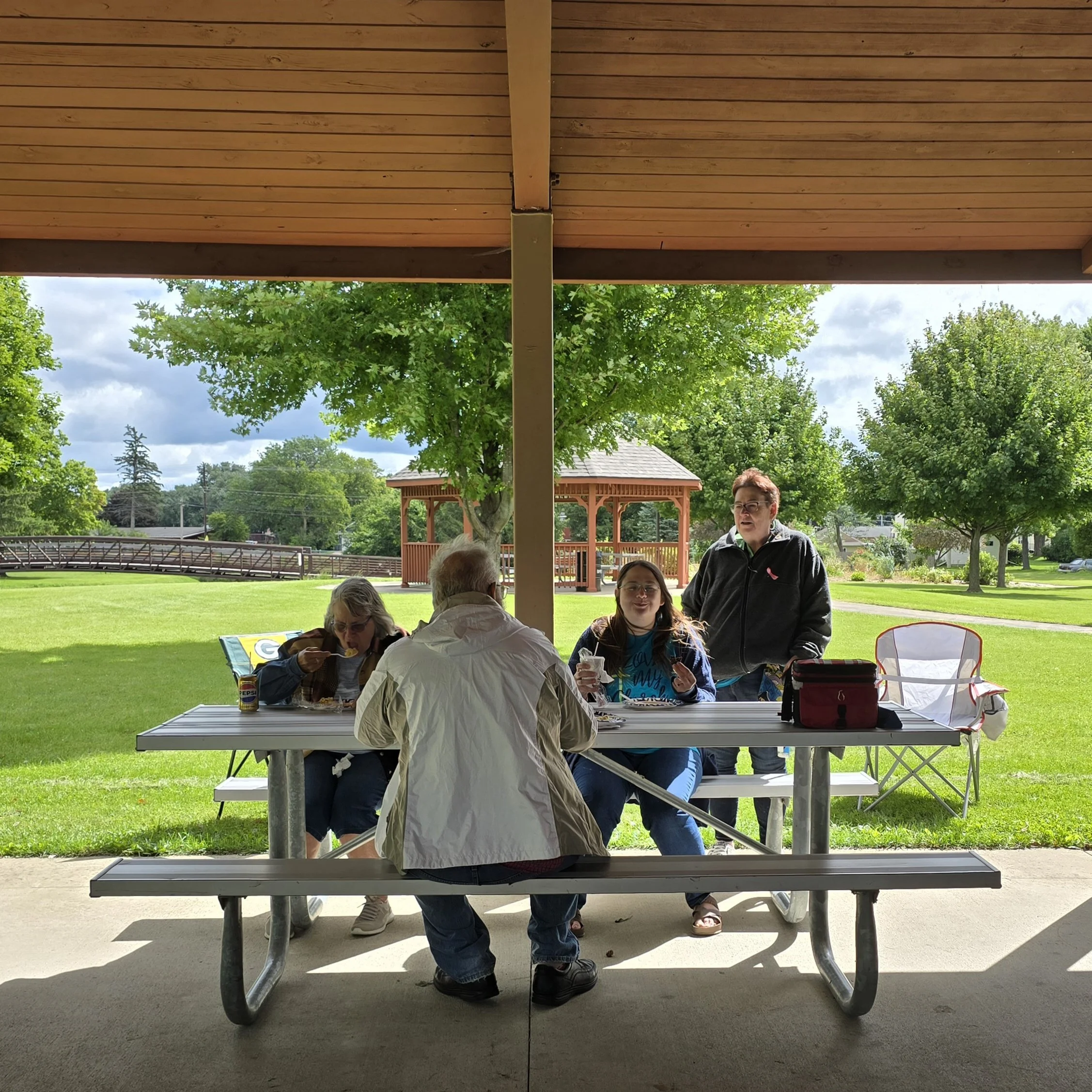 People sitting and standing around a picnic table outdoors under a shelter, with trees and a small pavilion in the background, on a cloudy day.