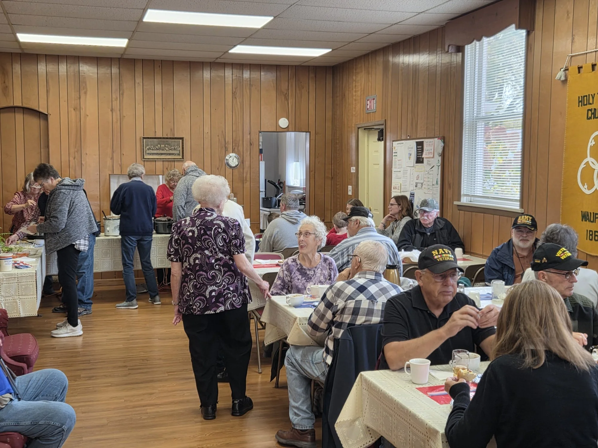A group of elderly people gathered in a dining hall with wood-paneled walls, some sitting at tables with beige tablecloths and others standing and serving food. There are windows with blinds and a bulletin board on the wall.