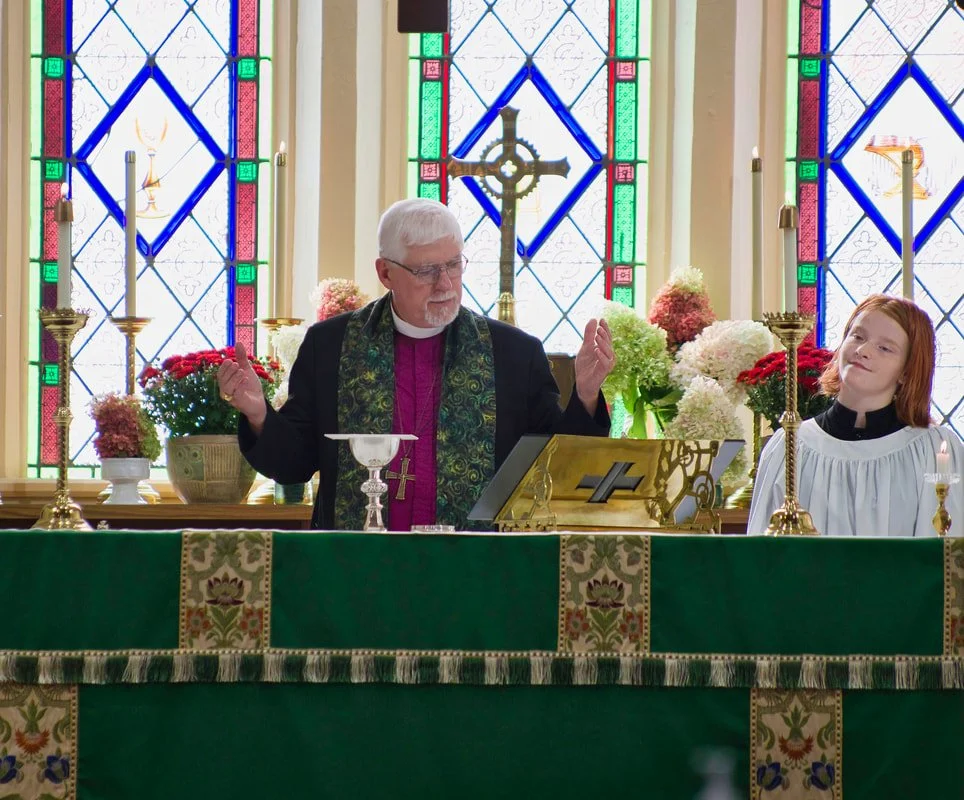 A church altar with a priest and an altar server during a service, featuring stained glass windows, candles, and floral arrangements.