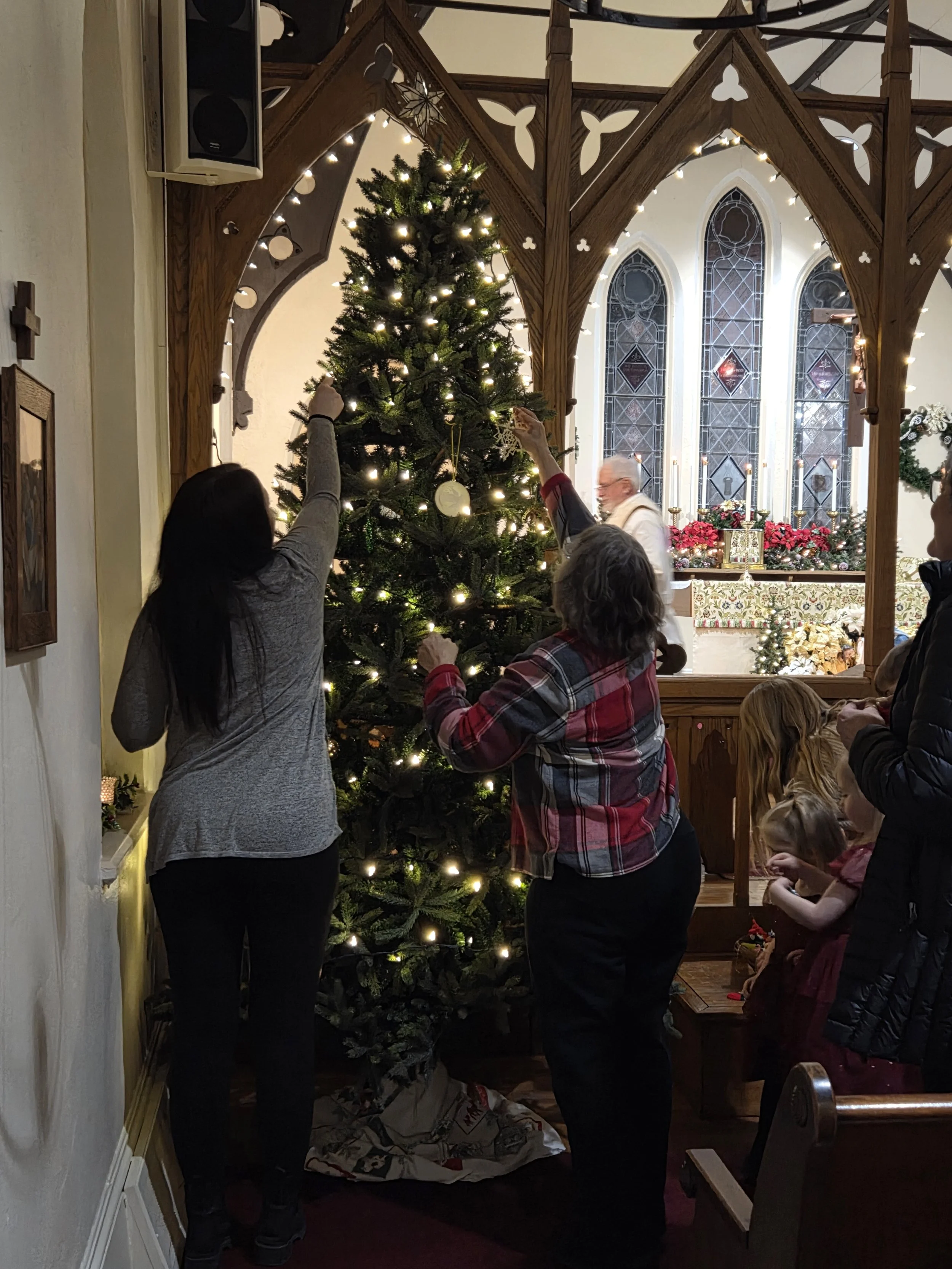 People decorating a Christmas tree inside a church or chapel with stained glass windows and wooden architecture.