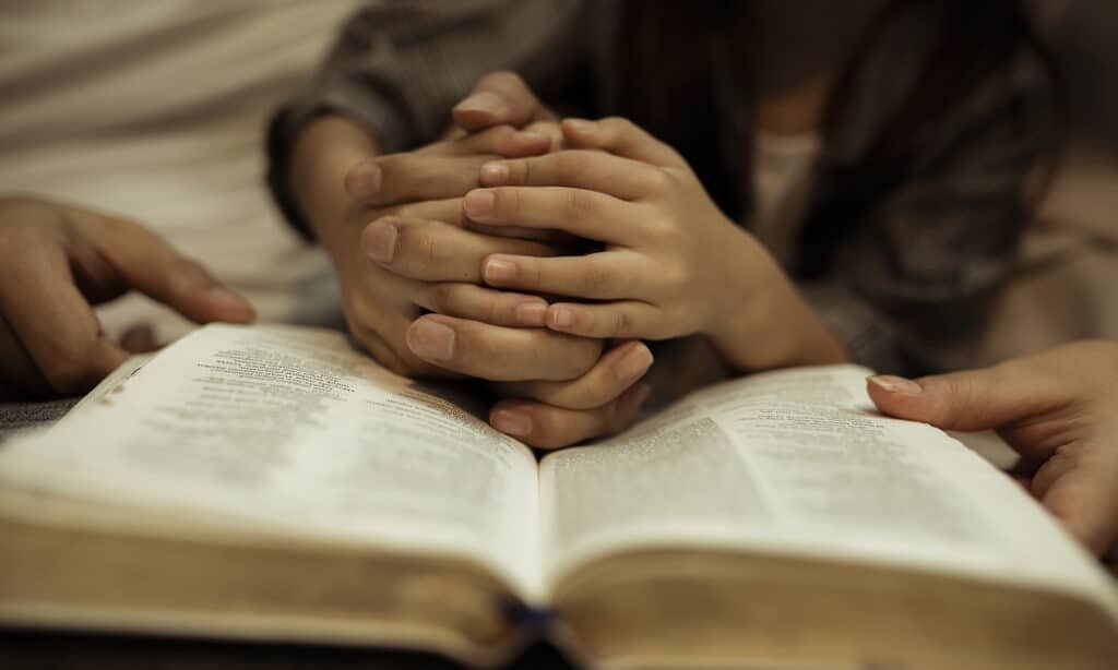 Three people have their hands clasped together over an open book, with two hands visible on each side, on a bed.