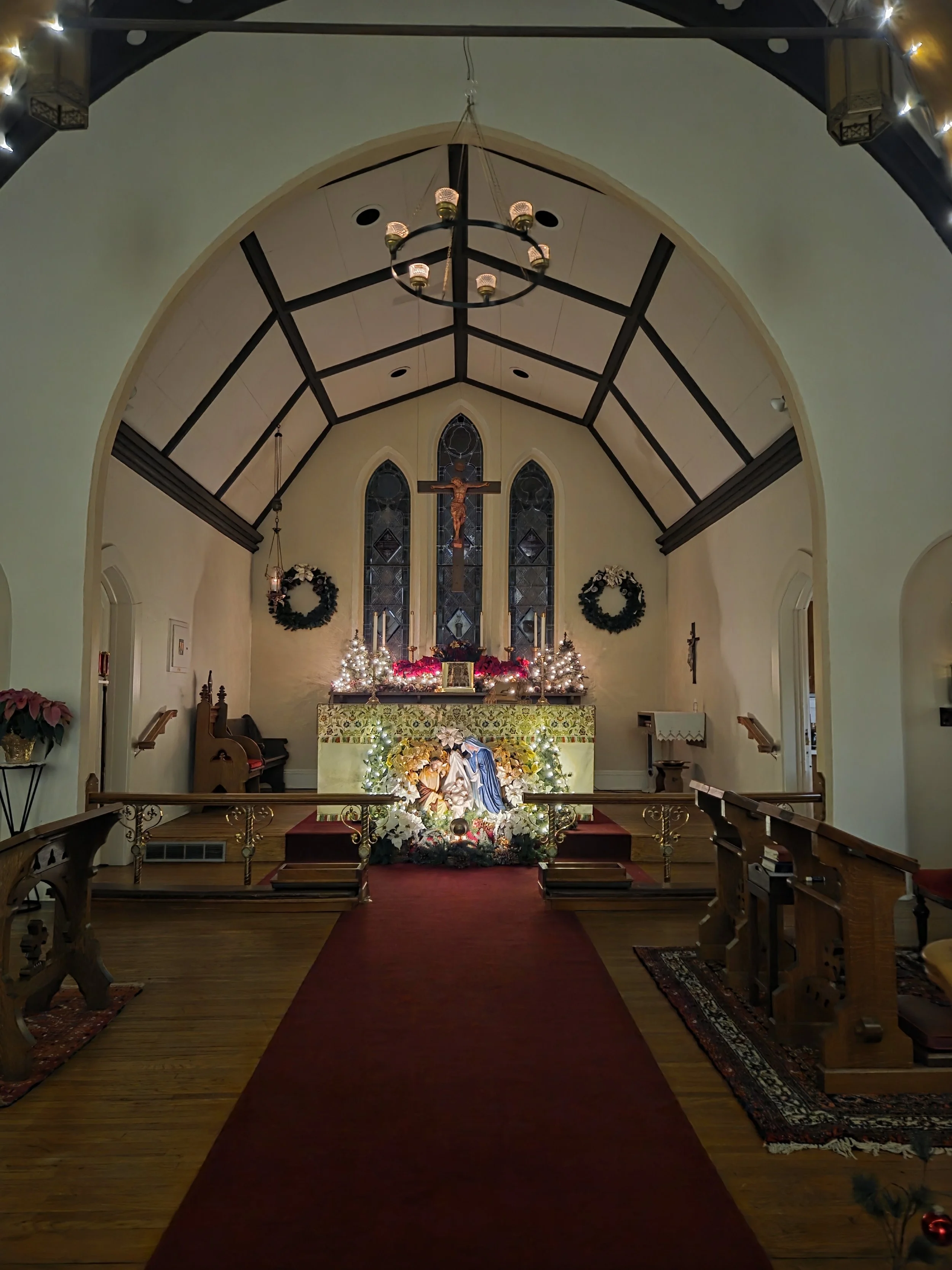 The interior of a church decorated for Christmas with a nativity scene in the foreground, Christmas wreaths on the wall, and candles on the altar. The church features stained glass windows and a prominent crucifix above the altar.