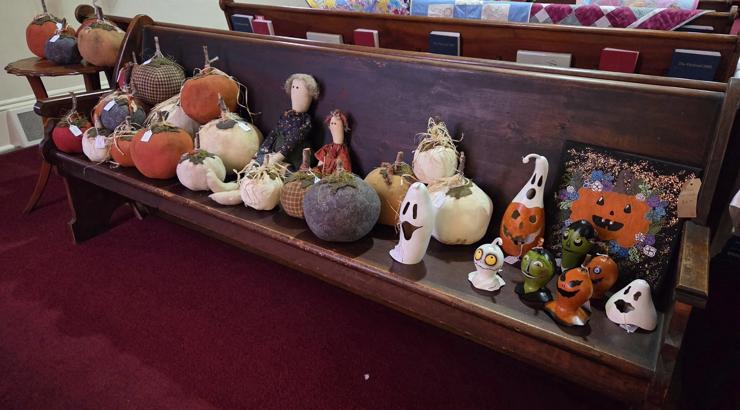 Display of decorative pumpkins, dolls, and Halloween-themed figurines on a wooden church pew with hymn books and a quilted blanket in the background.