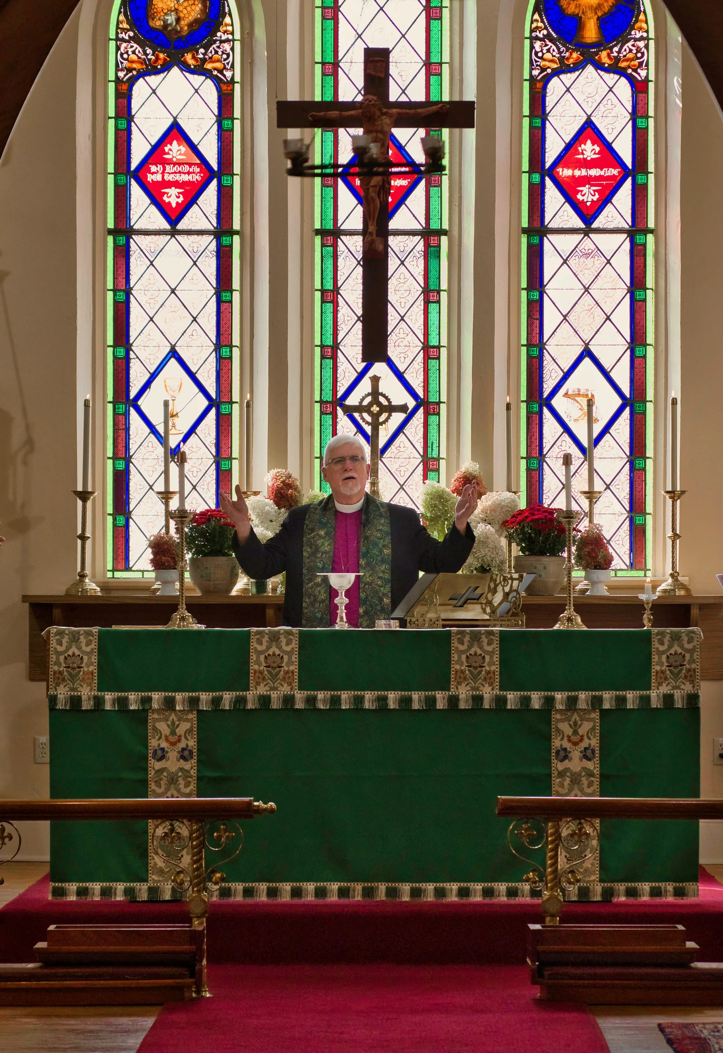 A priest standing at the altar of a church, with arms raised, in front of three stained glass windows. The altar is decorated with white and red flowers, candles, and a green altar cloth. A crucifix hangs above the altar.