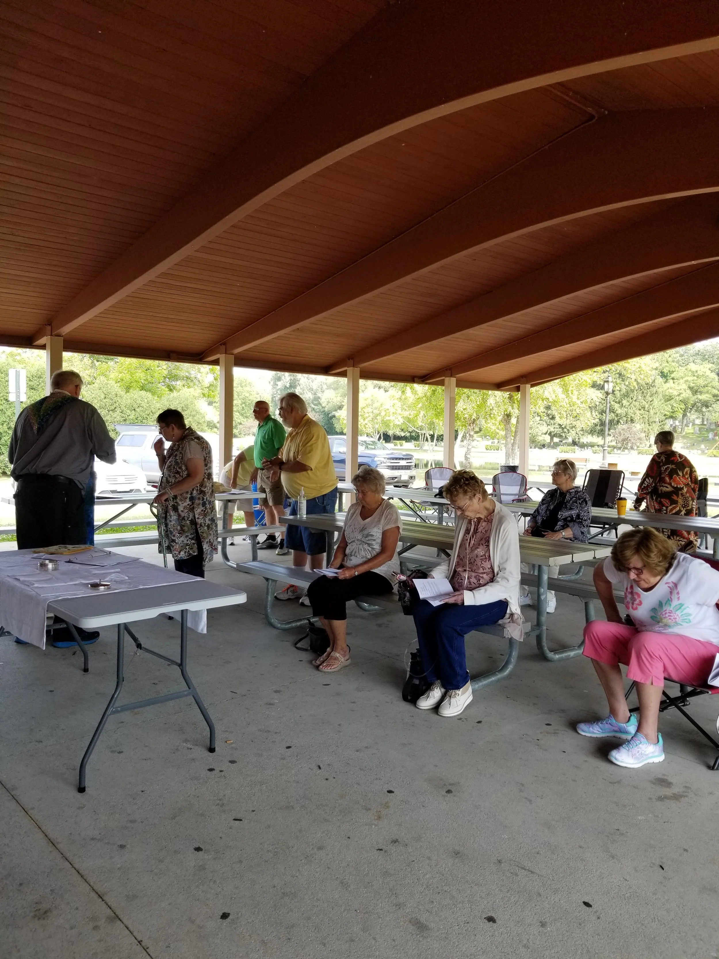 A group of people sitting and standing under a covered outdoor pavilion, reading and engaging in activities.