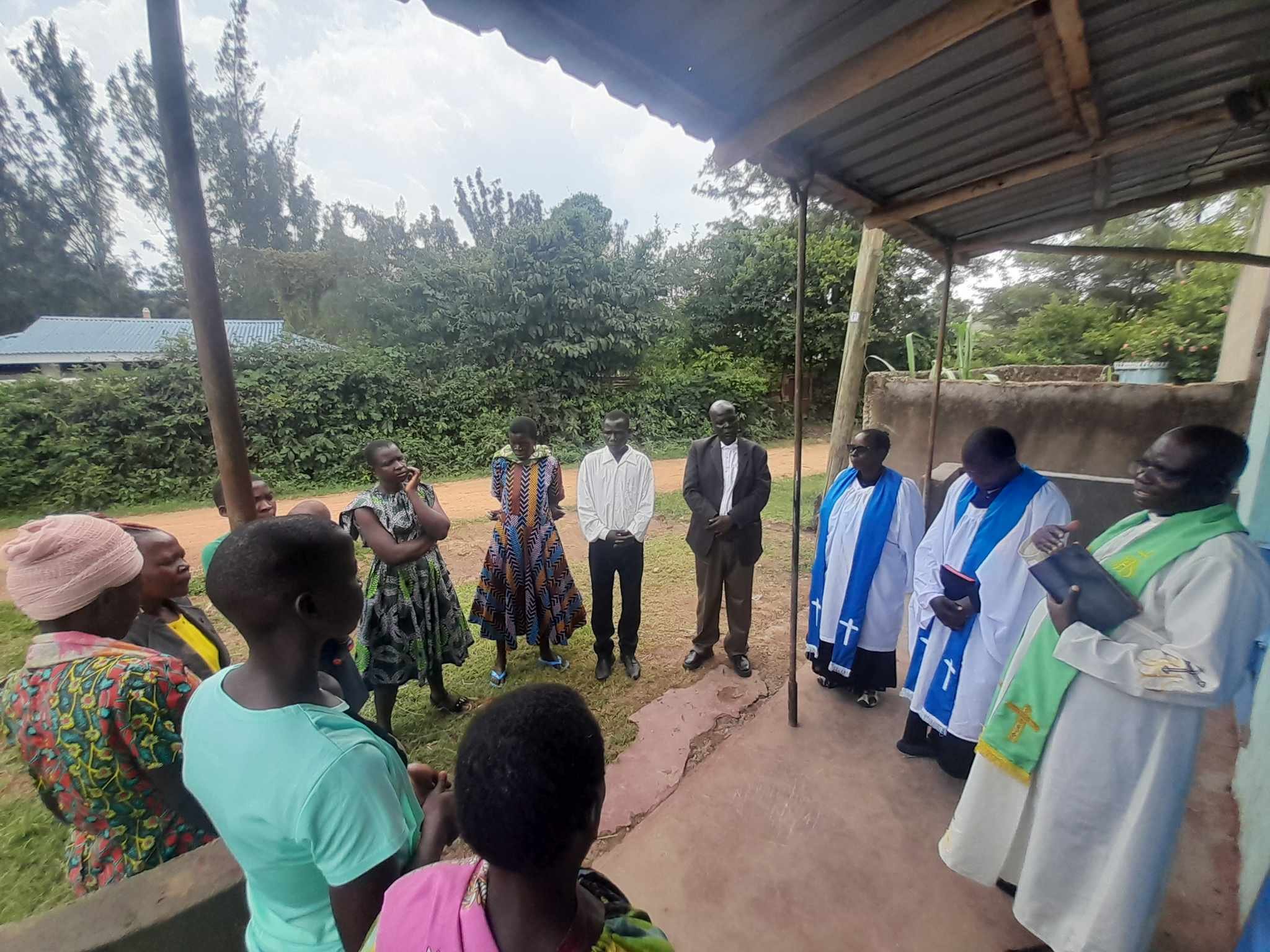 A group of people standing outdoors in a circle under a makeshift shelter, engaging in a discussion or meeting. The background shows greenery and trees.