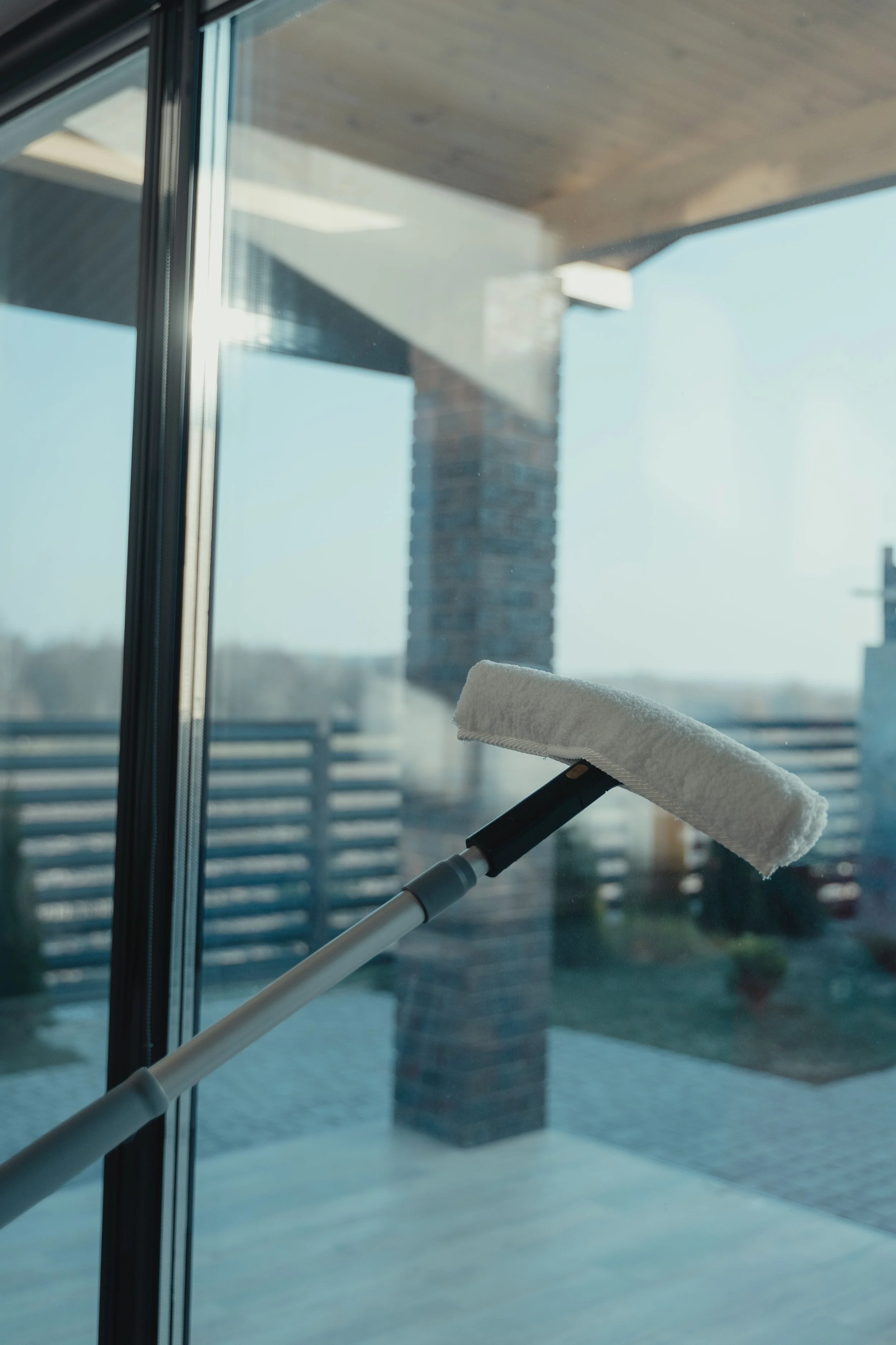 A cleaning spray mop with a white cloth head cleaning a glass window, with a modern building and blue sky visible through the window in the background.