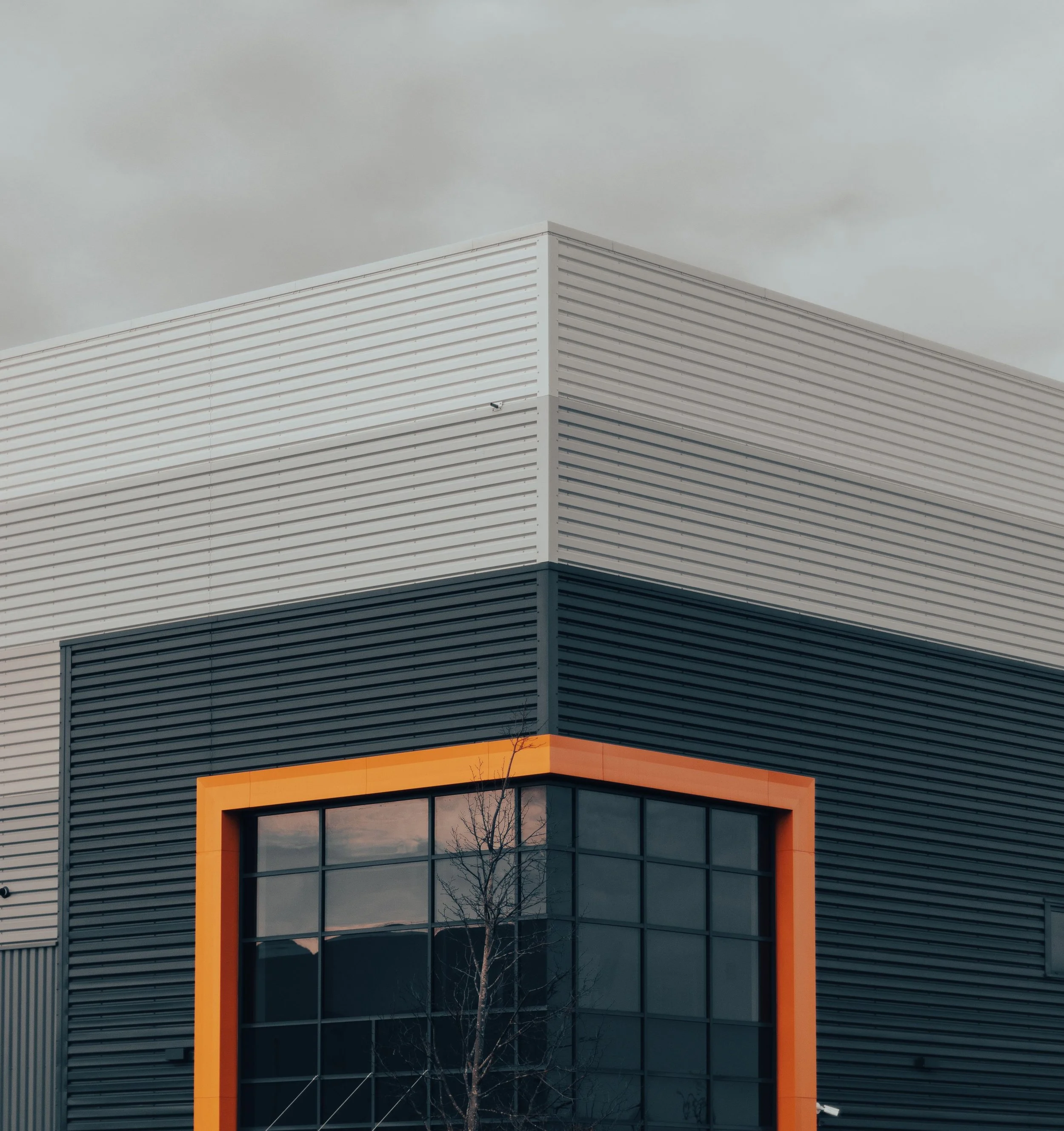 Modern building with large glass windows, gray horizontal siding, and an orange-frame balcony or architectural feature, with a leafless tree in front and overcast sky in the background.