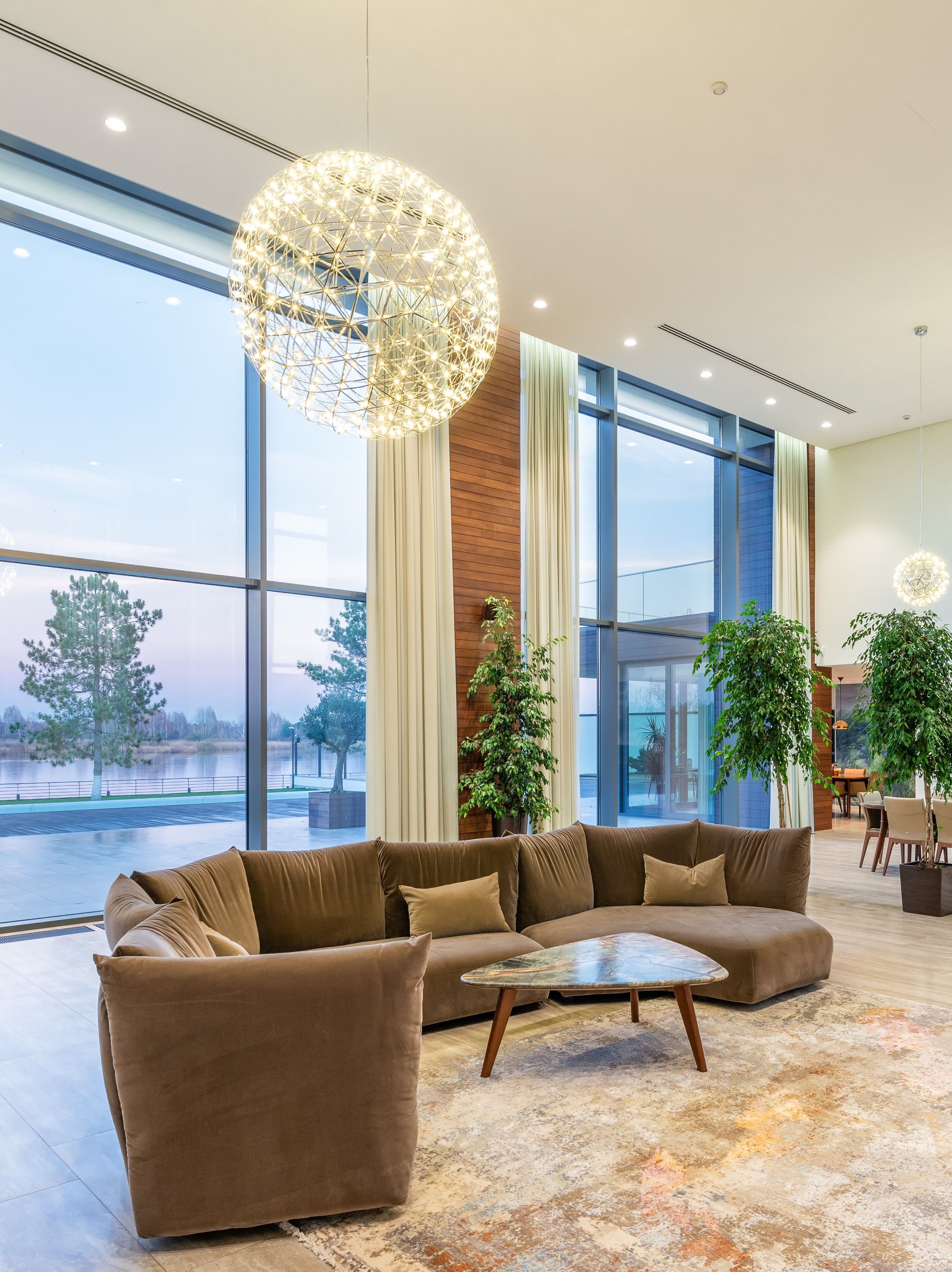 Modern indoor lounge area with large floor-to-ceiling windows, beige sectional sofa, marble coffee table, and decorative plants.