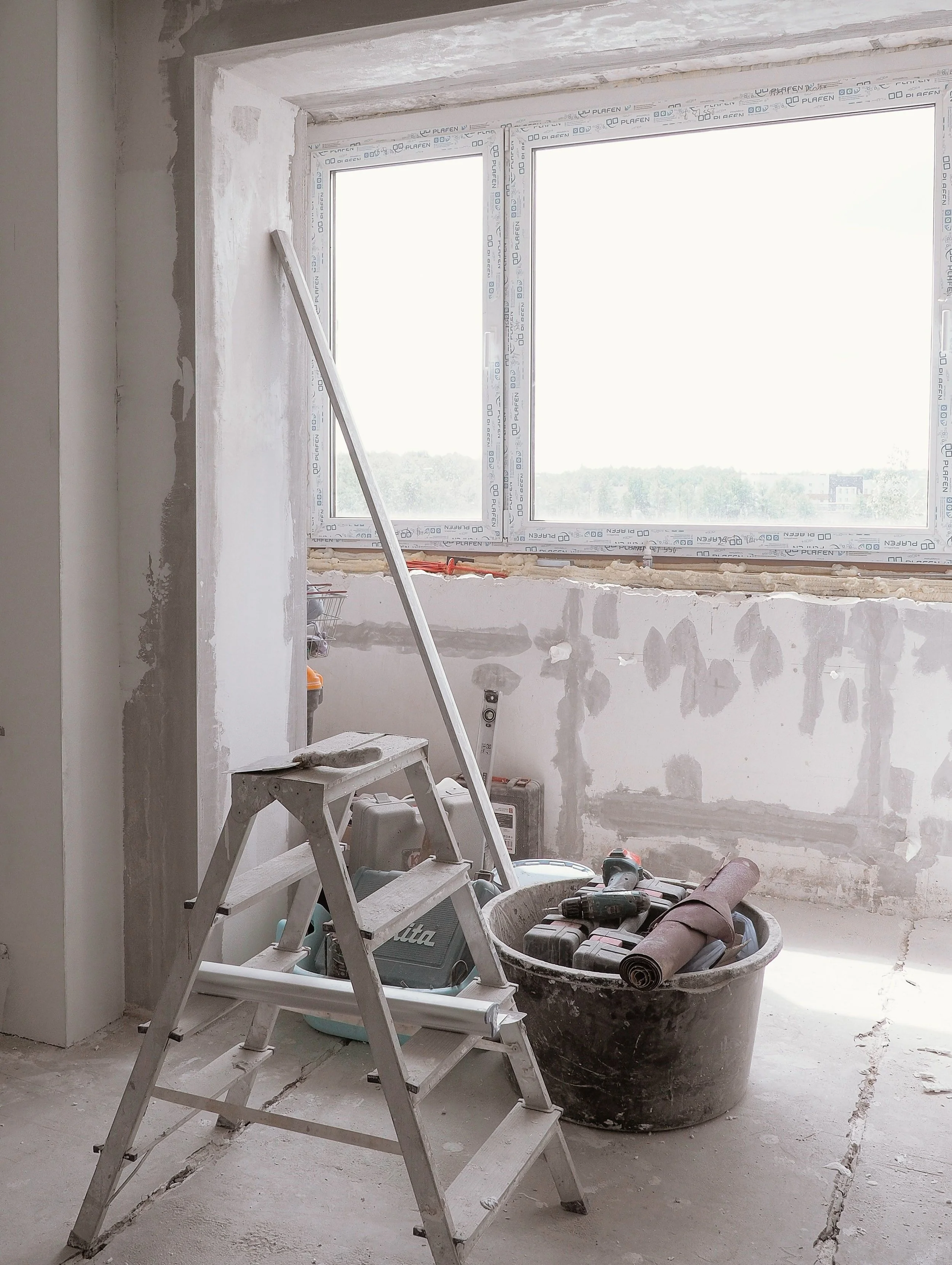 An unfinished room under construction with large window, construction tools, ladder, bucket, and rolled materials on the floor.
