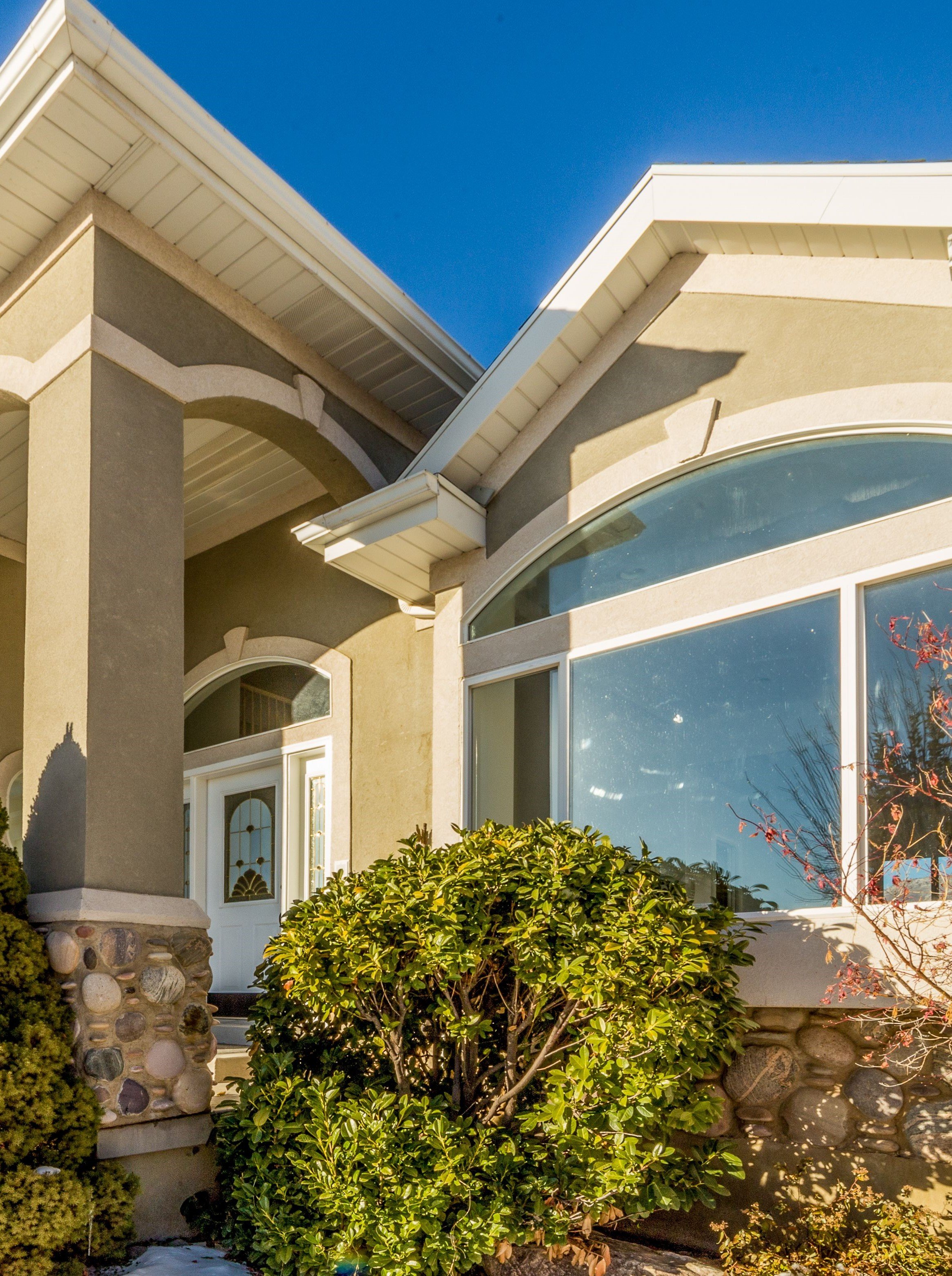 Close-up view of a house's exterior with beige stucco walls, white trim, large curved window, and greenery including bushes and trees in front, under a clear blue sky.